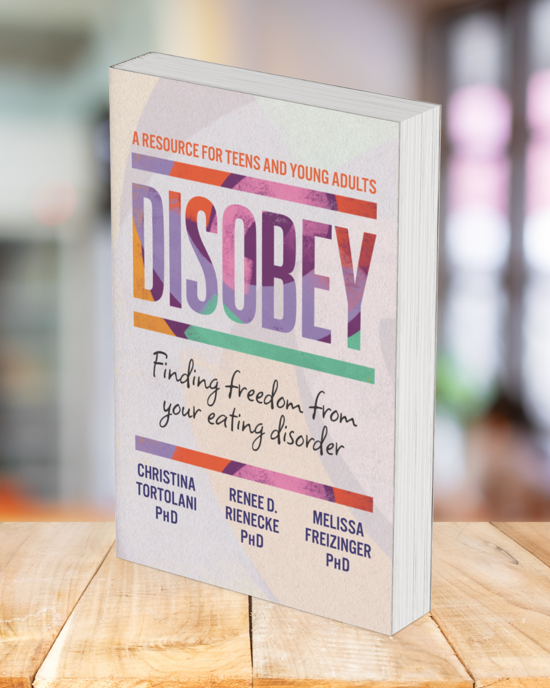Book titled 'Disobey: Finding Freedom from Your Eating Disorder,' by Christina Tortalani, Renee D. Rienecke, and Melissa Freizinger, with the subtitle 'A resource for teens and young adults,' standing on a wooden surface in a blurred indoor setting.