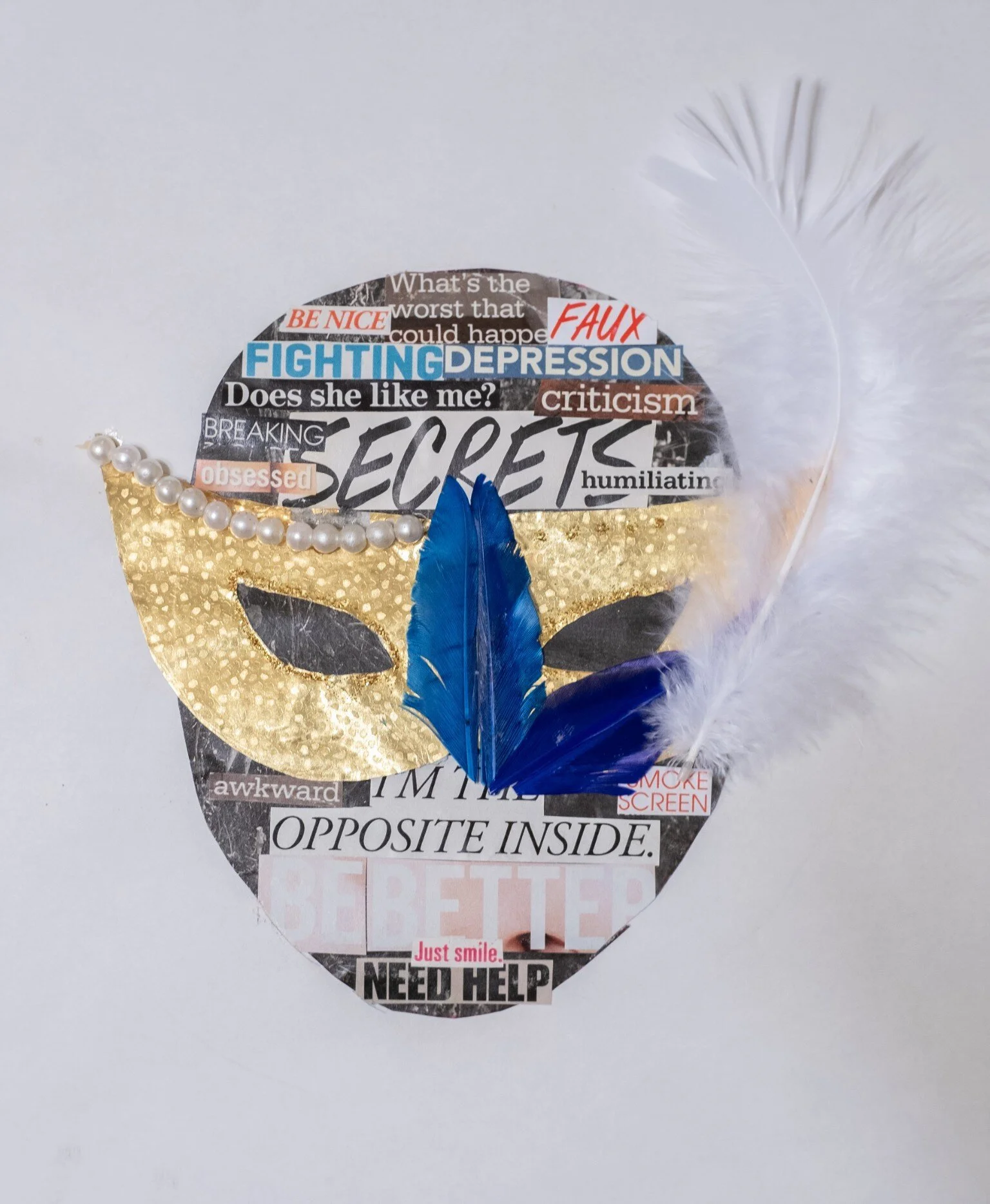 A collage in the shape of a face mask made from magazine cutouts with various words and phrases. The mask features a gold crown with pearl embellishments, and a cluster of blue and purple feathers attached at the bottom. The background is plain white.