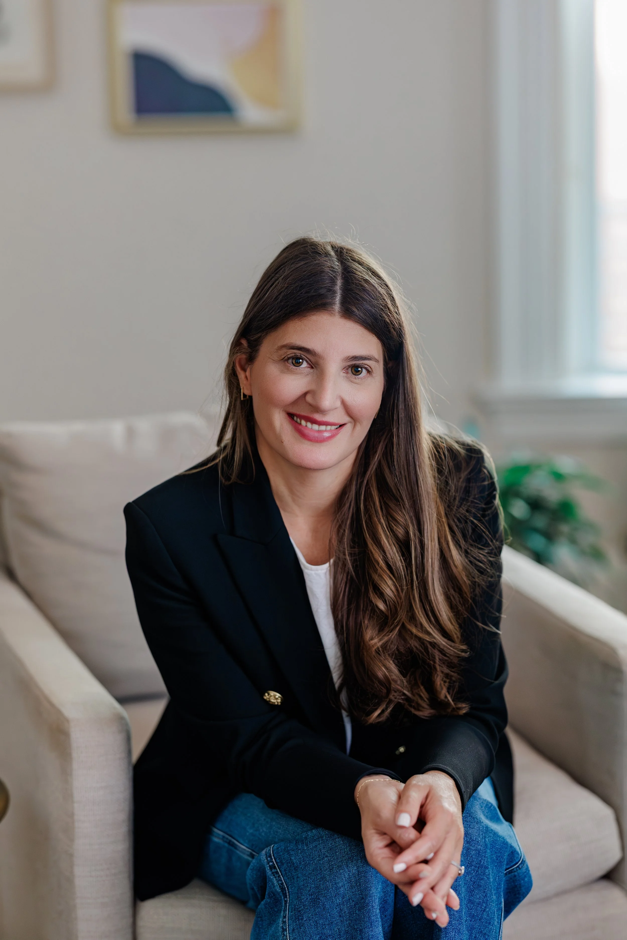 A woman with long brown hair, wearing a black blazer and blue jeans, sitting on a cream-colored couch indoors. She is smiling and looking at the camera with her hands clasped in her lap. There are framed artworks on the wall behind her and a window letting in light.
