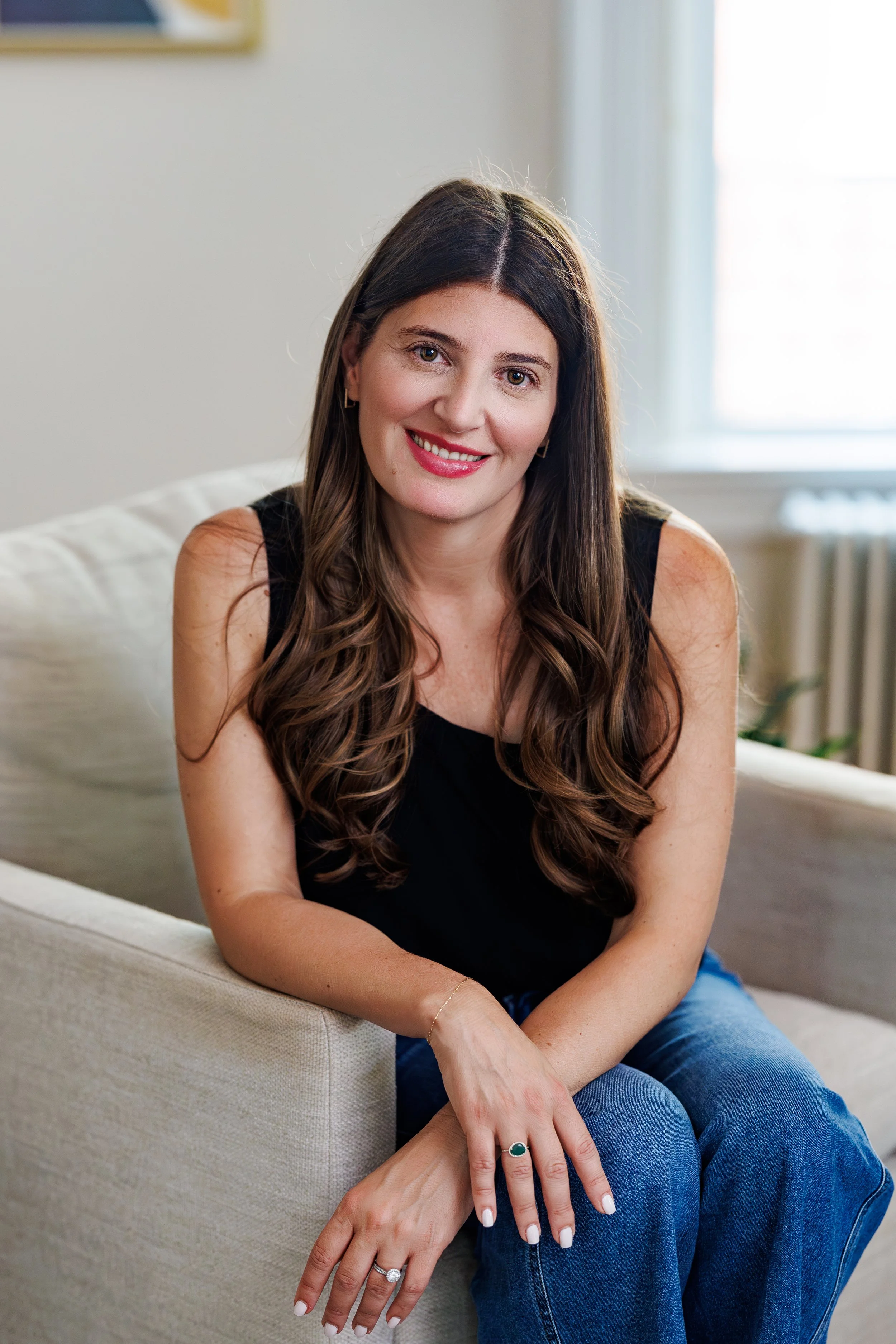A woman with long brown hair and light skin sitting on a beige sofa, smiling, wearing a black sleeveless top and blue jeans, with a window and white curtains in the background.