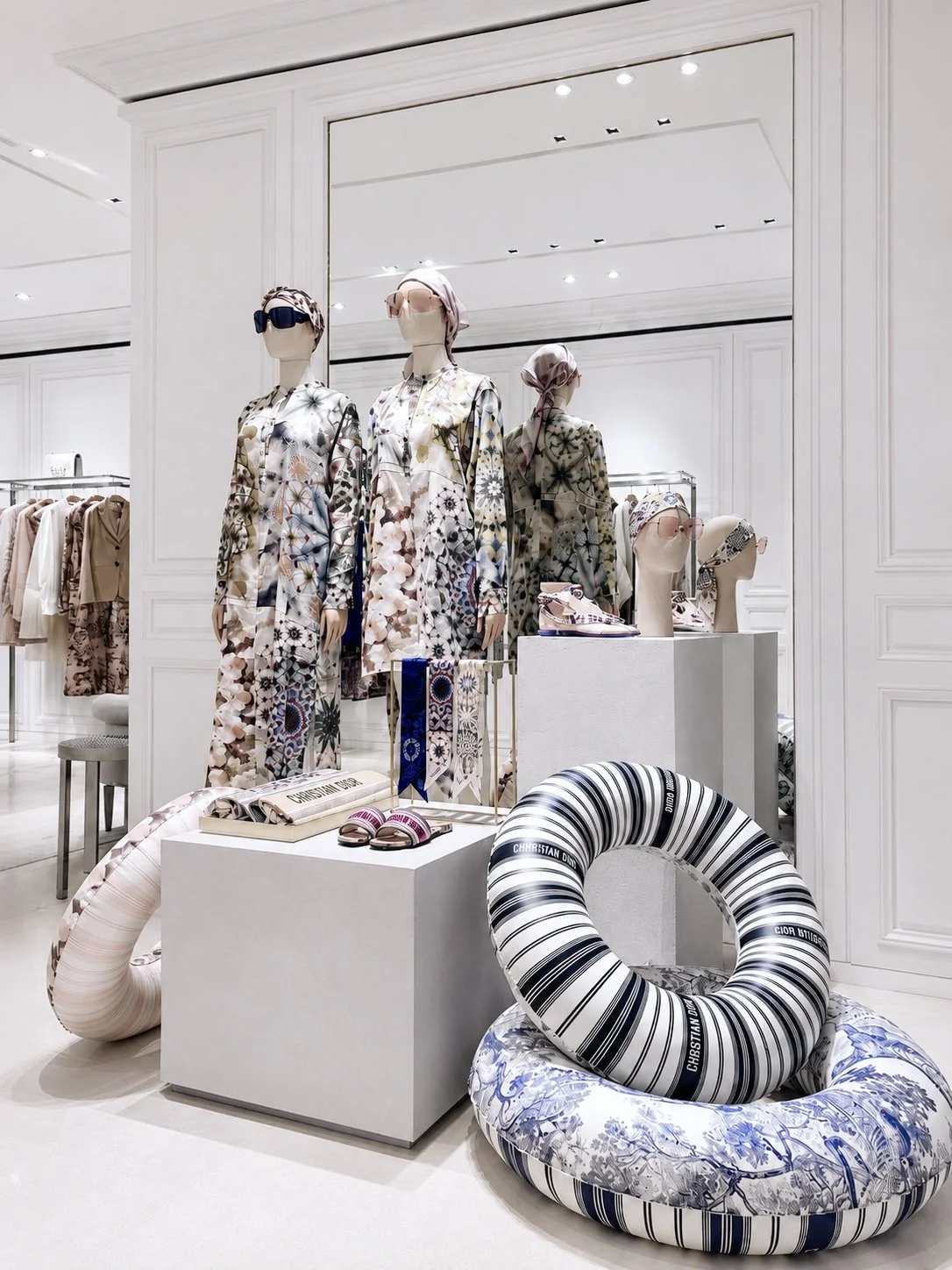 Vitrine de mode avec mannequins portant des robes à motifs floraux et des accessoires de soleil, décor blanc avec coussins en forme de bouée.