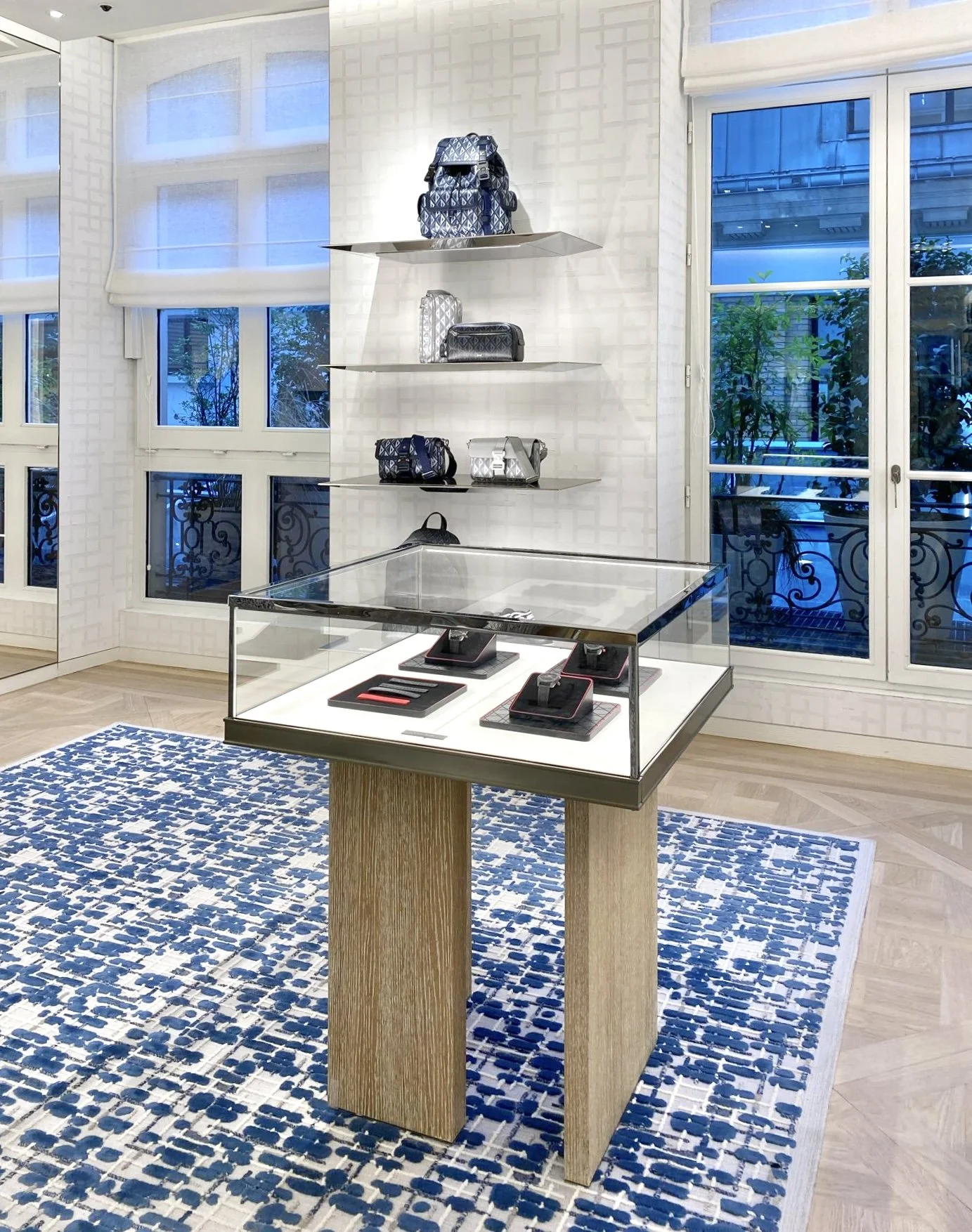 Intérieur d'une boutique de luxe avec des sacs à main exposés sur des étagères blanches et un présentoir en verre avec des montres. La pièce a de grandes fenêtres avec des volets blancs et un tapis bleu et blanc au sol.