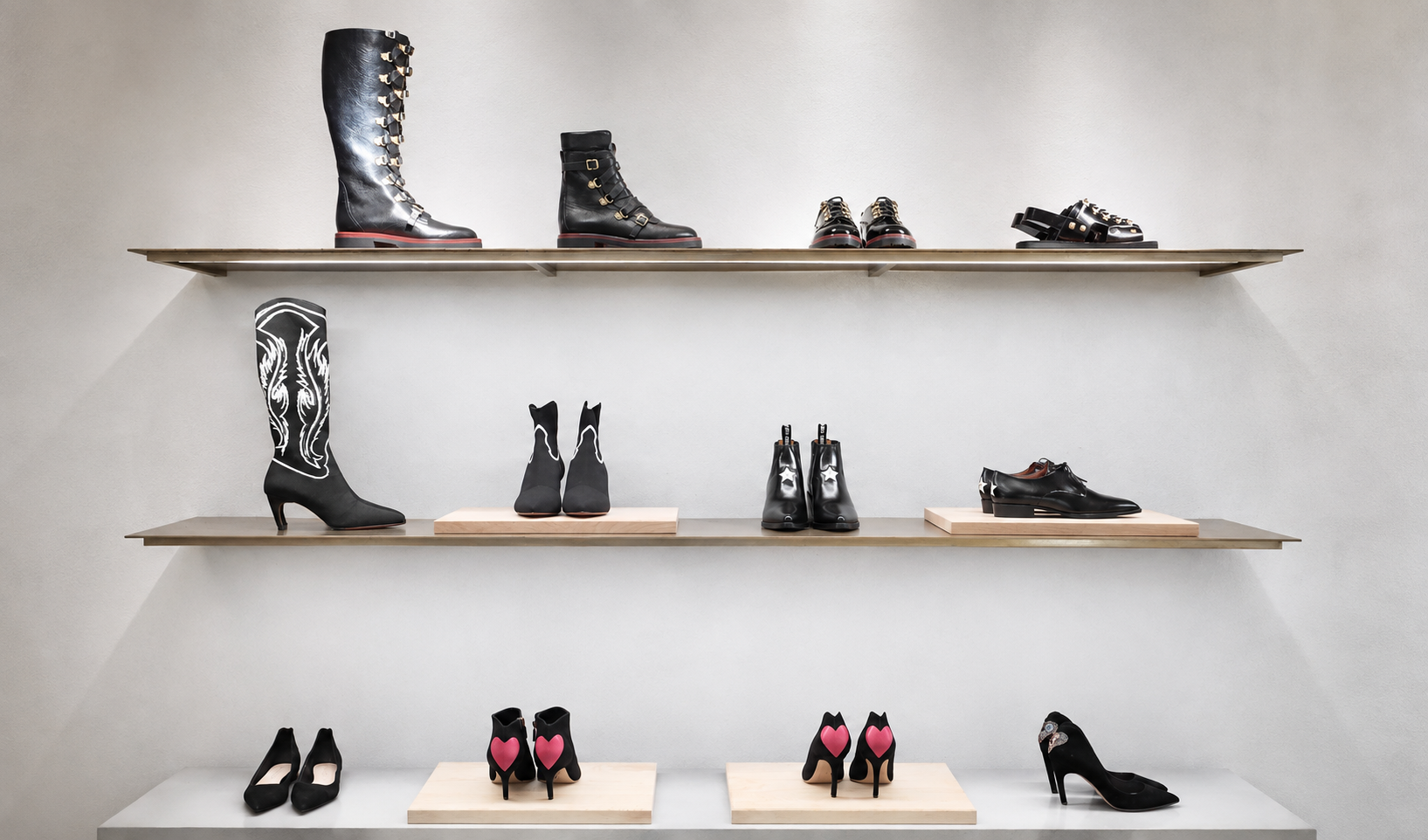Étagères avec différentes chaussures à talons et bottes, dont des bottes noires avec des détails blancs, des chaussures à talons avec de cœurs roses, et des chaussures noires classiques, dans un magasin de chaussures.
