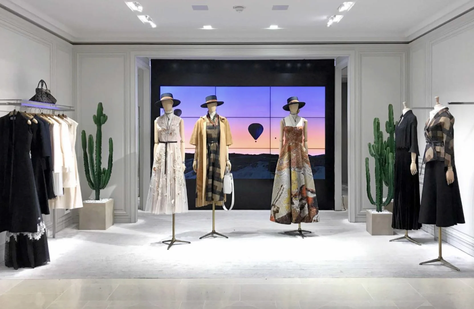 Vitrine d'une boutique de vêtements avec mannequins portant des robes d'été, chapeaux, et sacs à main. Décorée avec des cactus artificiels et un mur avec écran affichant une image de montgolfière dans le ciel au coucher de soleil.