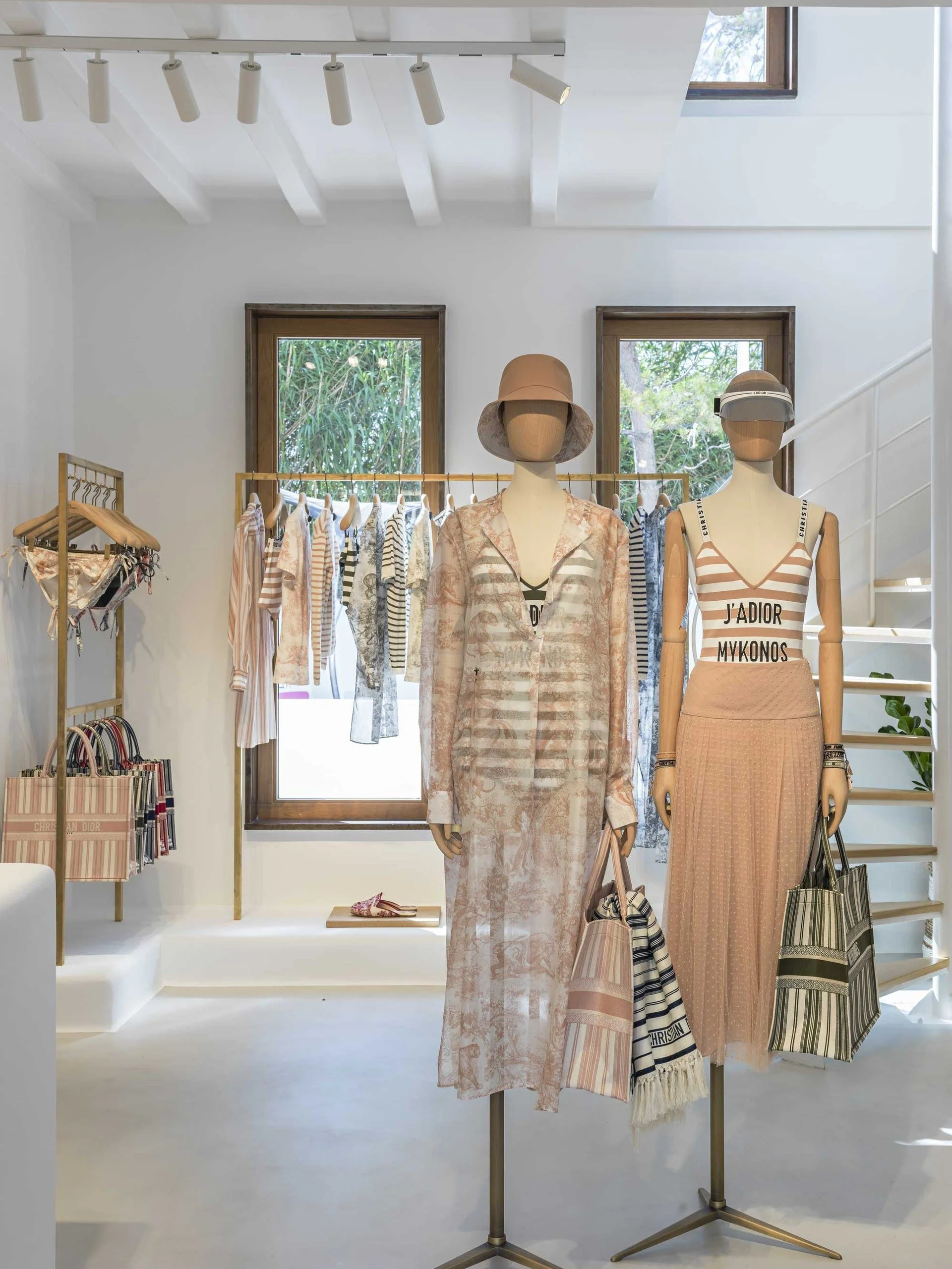 Vitrine de vêtements avec mannequins portant des tenues légères aux motifs floraux et rayés, écharpes et sacs à main, dans un magasin de mode.