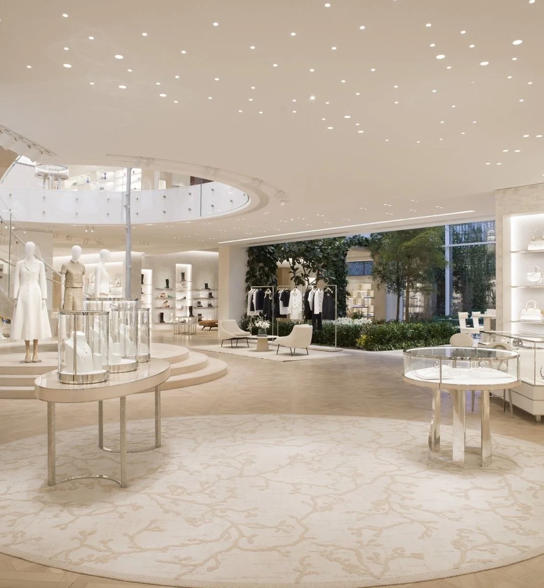 Intérieur d'une boutique de mode moderne avec des mannequins vêtus de vêtements blancs, des étagères avec des sacs et accessoires, et un espace d'accueil élégant avec des fauteuils et des arbres en intérieur.