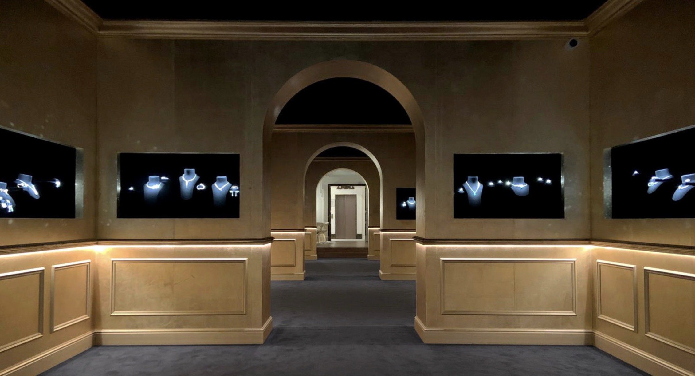 Une salle d'exposition avec des écrans affichant des bijoux en diamants, entourée de murs beige et pavée de tapis gris, avec un plafond haut et une architecture à arches.