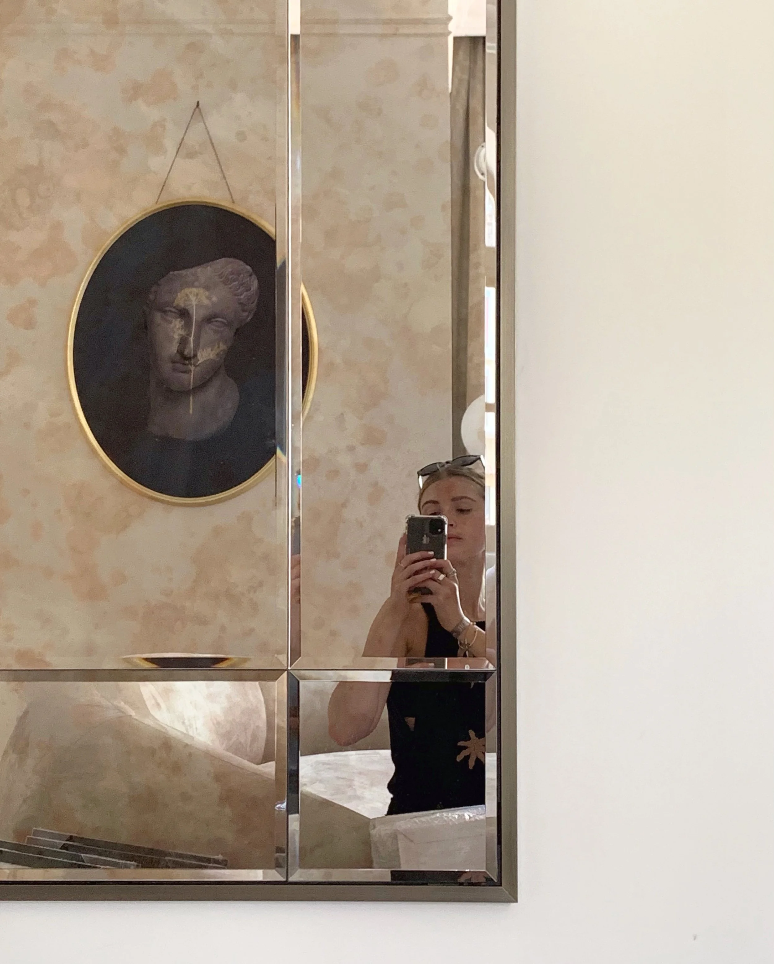 Reflet d’un jeune femme prenant une photo avec son téléphone dans un miroir en miroir. À côté, un tableau avec une sculpture de tête ancienne en relief.