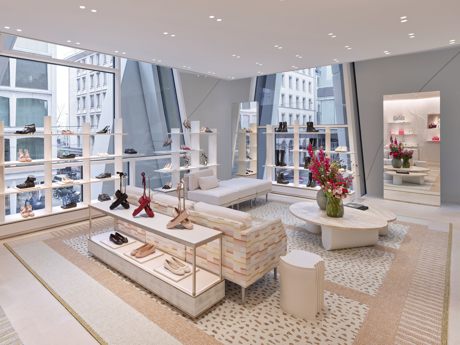 Vitrine d'une boutique de chaussures avec des chaussures sur des étagères, un canapé blanc, une table en marbre avec des vases de fleurs, un miroir mural, et un tapis beige et blanc.