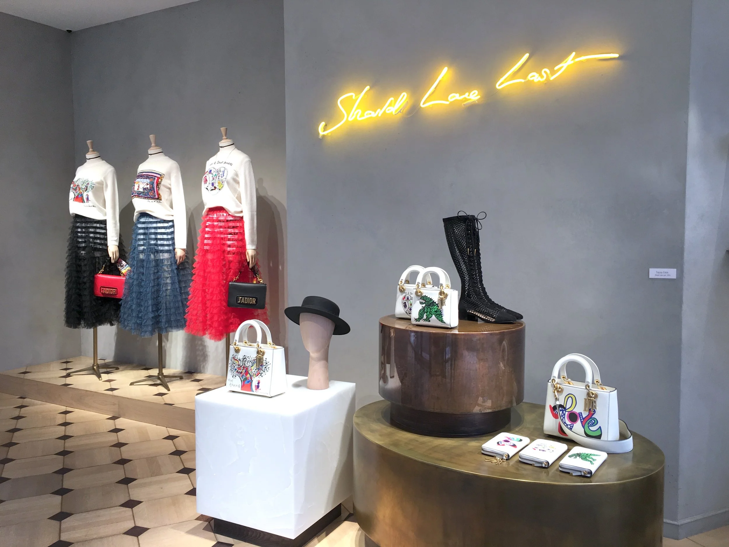 Vitrine de vêtements de mode avec trois mannequins portant des jupes longues à volants, une enseigne lumineuse jaune avec le texte 'Should Love' sur le mur, et différentes sacs à main décoratifs colorés sur une table et un support. Le sol a un motif géométrique en bois clair et noir.