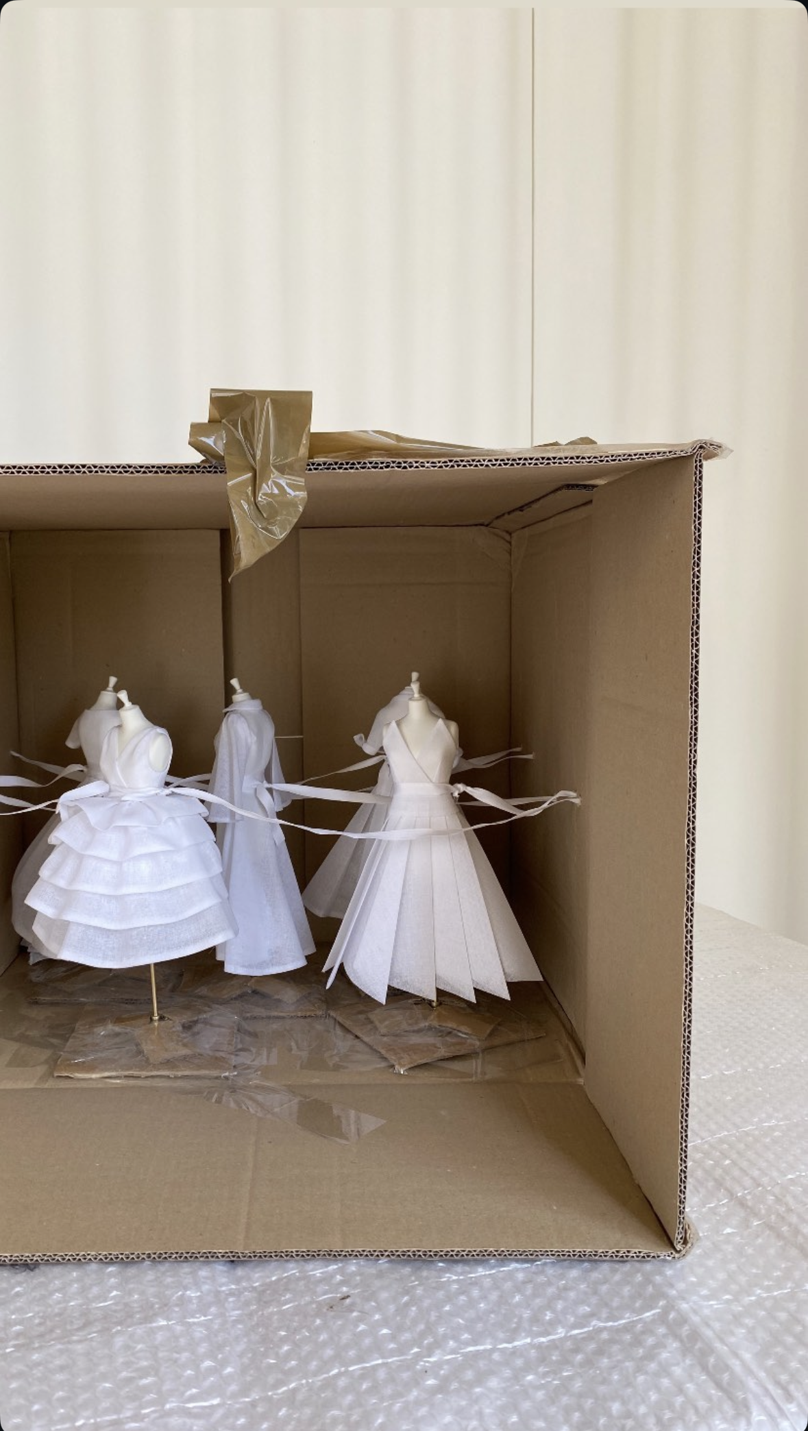 Une boîte en carton contenant quatre poupées mannequins en tissu blanc, représentant des robes de mariée ou des costumes, utilisées pour la présentation ou la décoration.