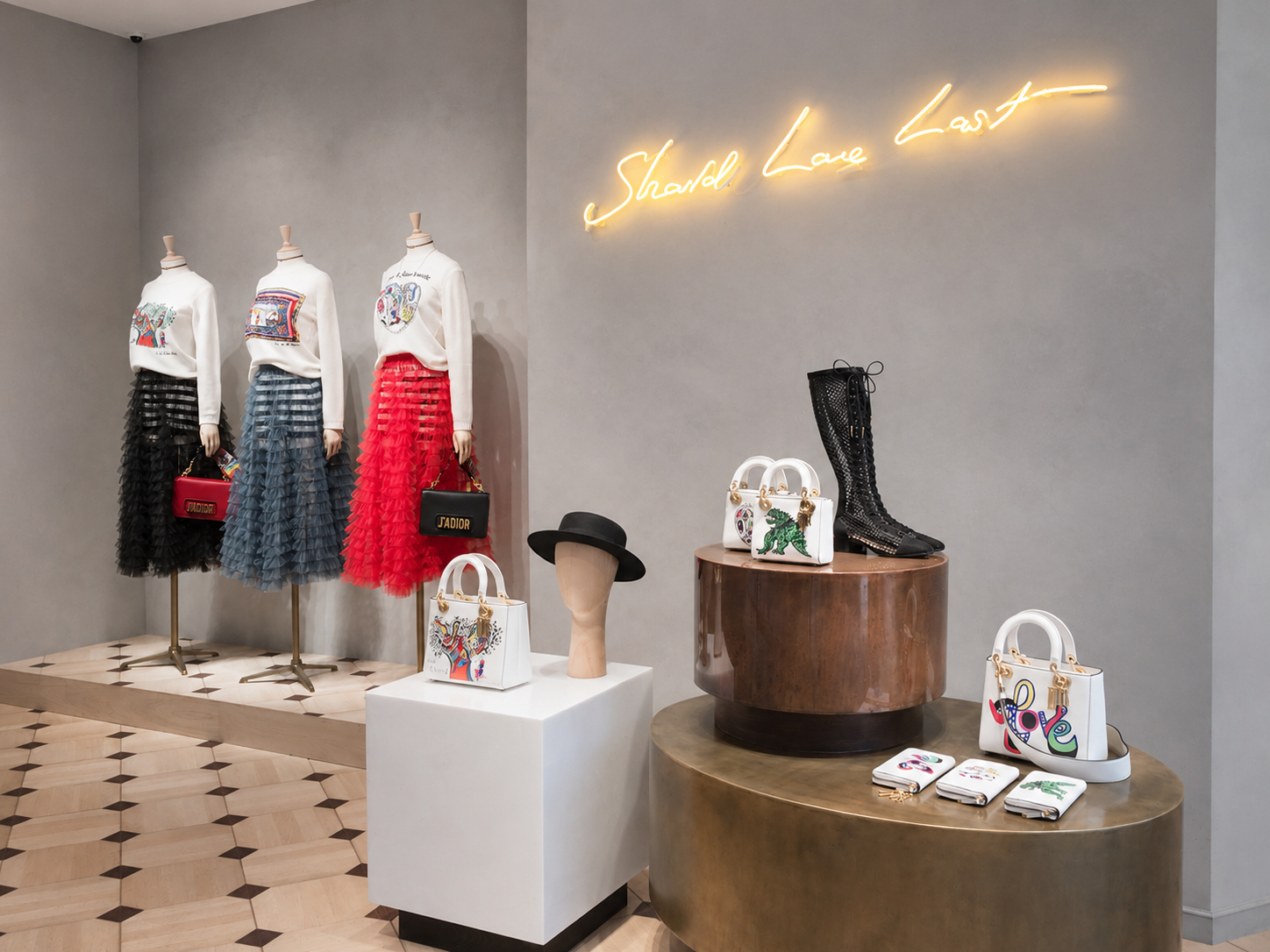 Vitrine de vêtements de mode avec trois mannequins portant des jupes en tulle roulé et des pulls imprimés, sacs à main colorés et bottes en cuir noir, décor moderne avec une enseigne en néon "Should Leave Last"