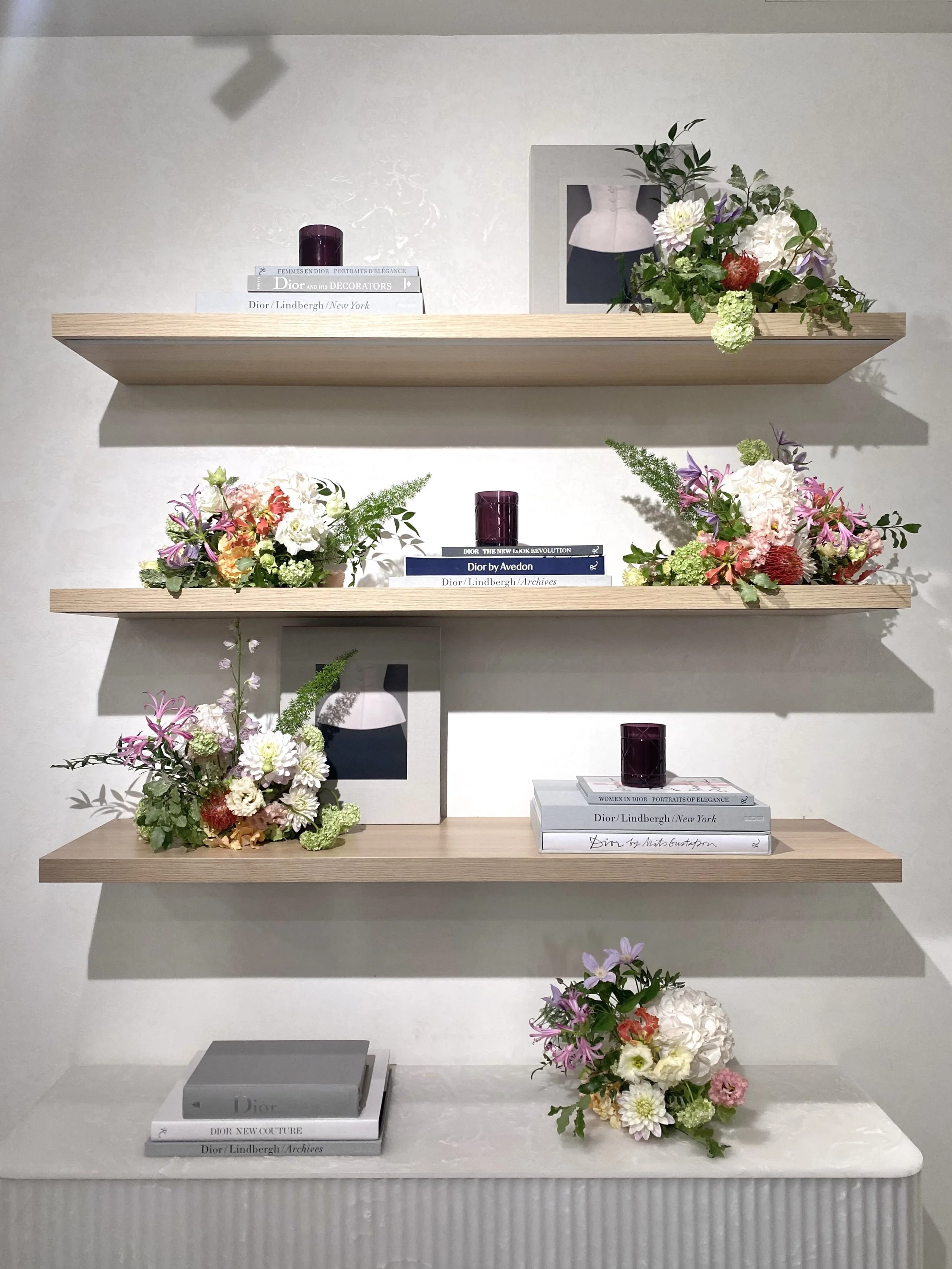 Trois étagères en bois aux décorations florales et livres, décoration intérieure moderne. La première étagère a un livre sur Dior, une bougie violette, et une photo en noir et blanc. La deuxième étagère a un arrangement floral coloré, avec un livre sur Dior et une bougie violette. La troisième étagère a un bouquet floral, aussi avec un livre sur Dior.