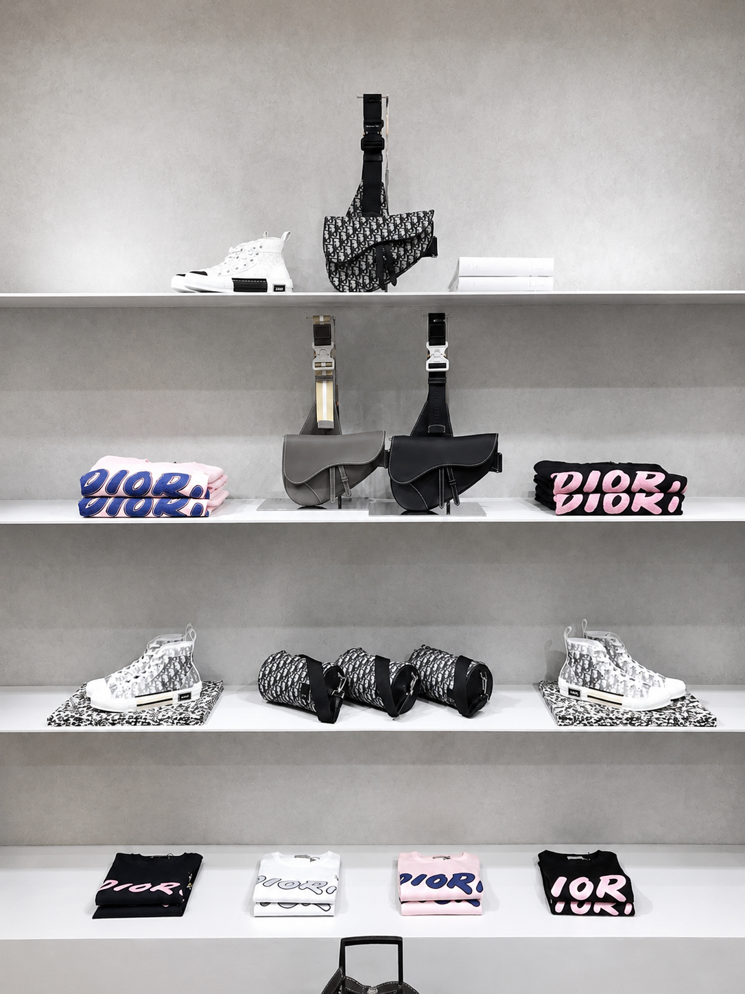Vitrine de mode avec des sacs, des baskets, des t-shirts et des accessoires Dior en noir, blanc, gris et rose.