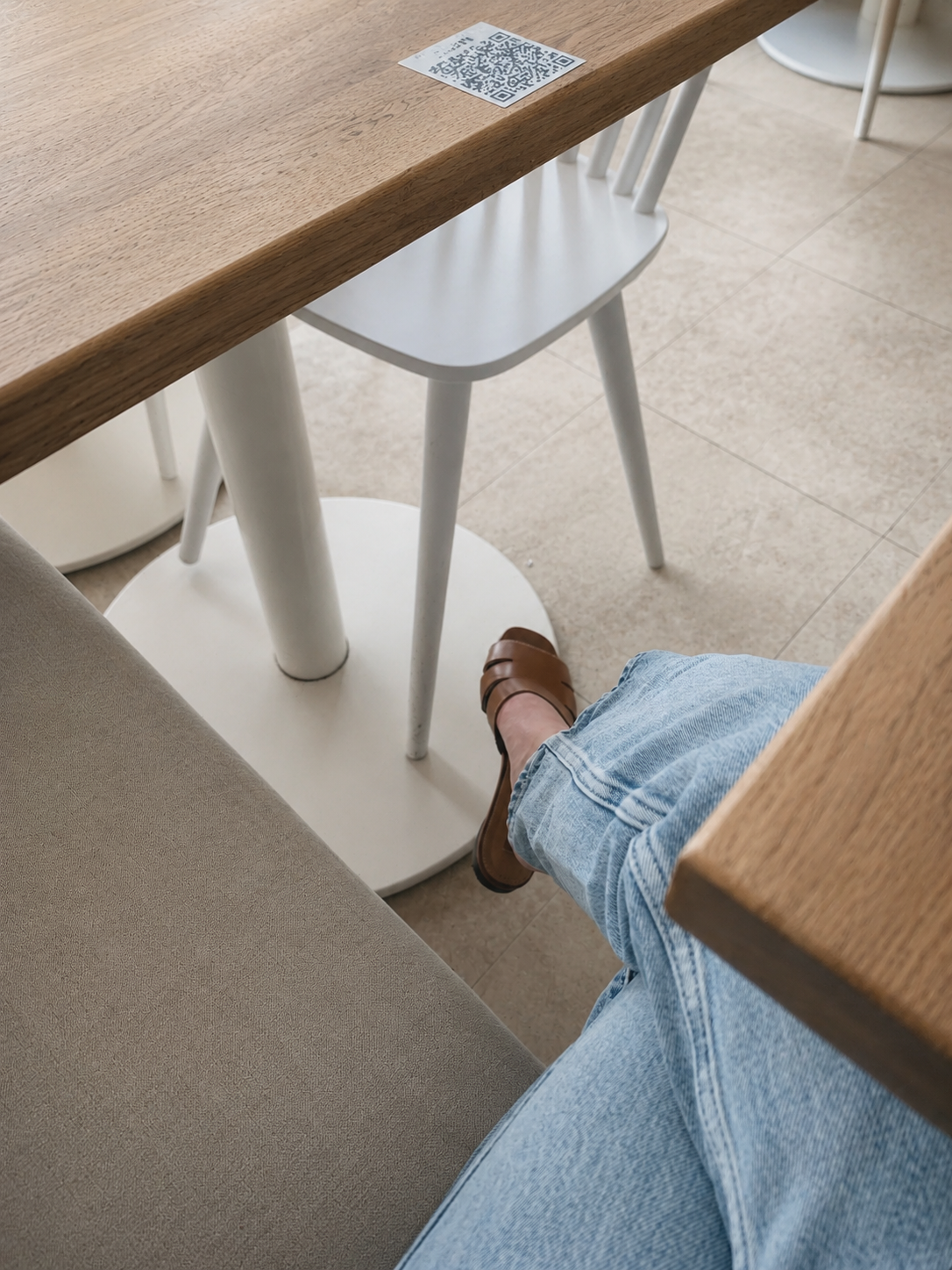 Une personne en jeans et sandales marron assise à une table en bois, avec un QR code visible sur la table.
