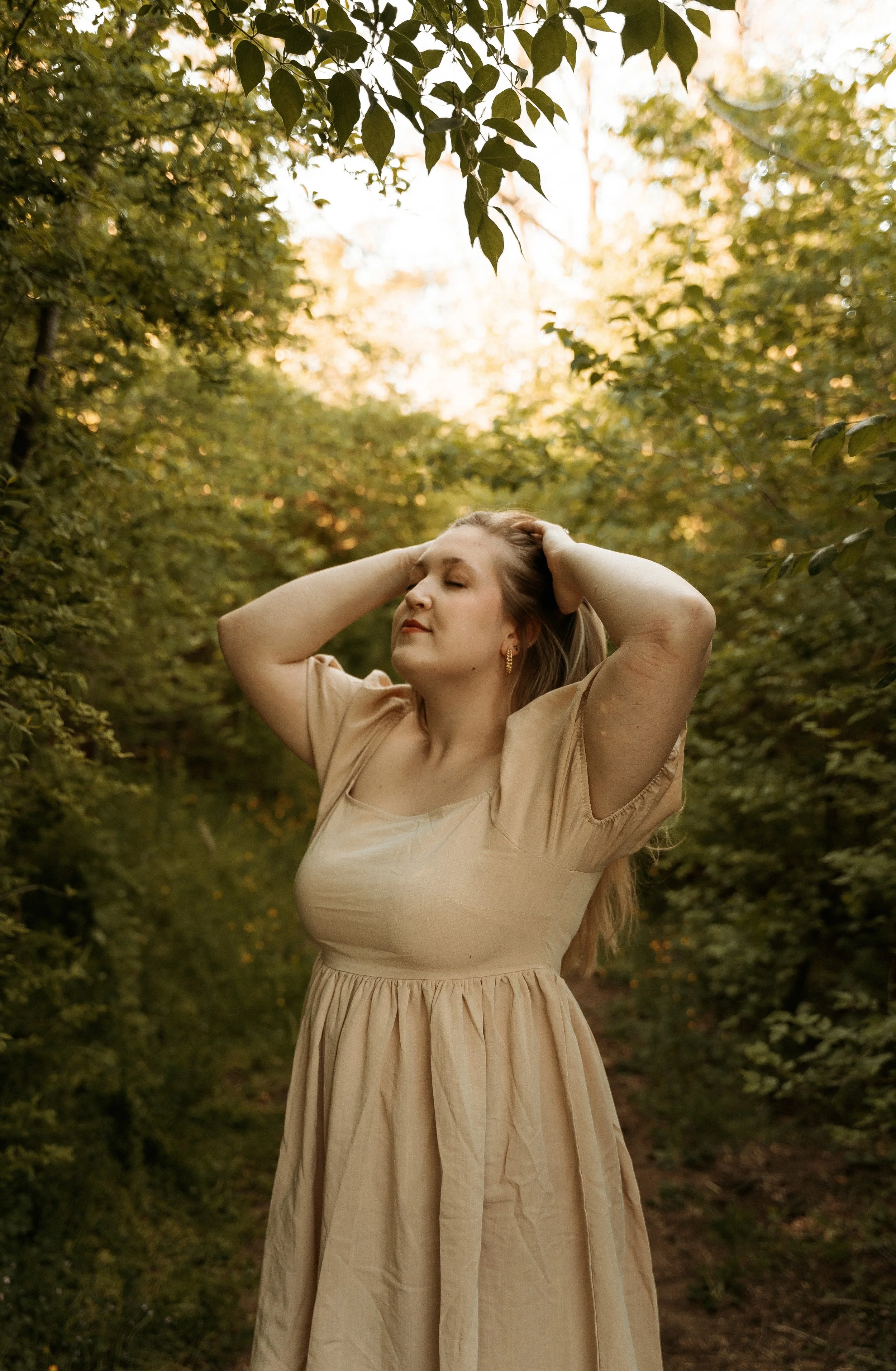woman stands in forest with eyes closed and her face tilted up towards the sky