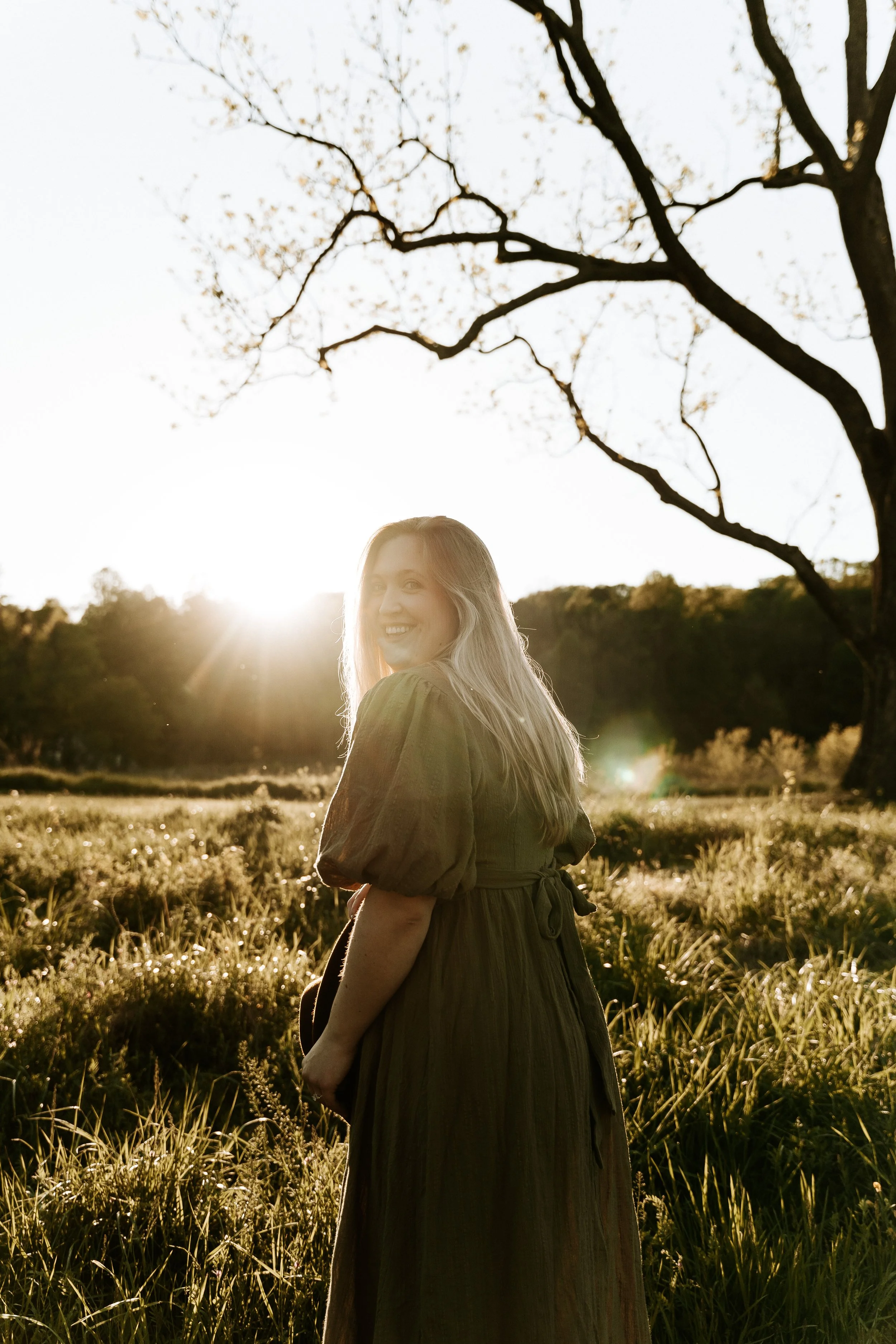 woman standing in a field wearing a green dress looks back and smiles towards the camera. you can see the sun shining behind her
