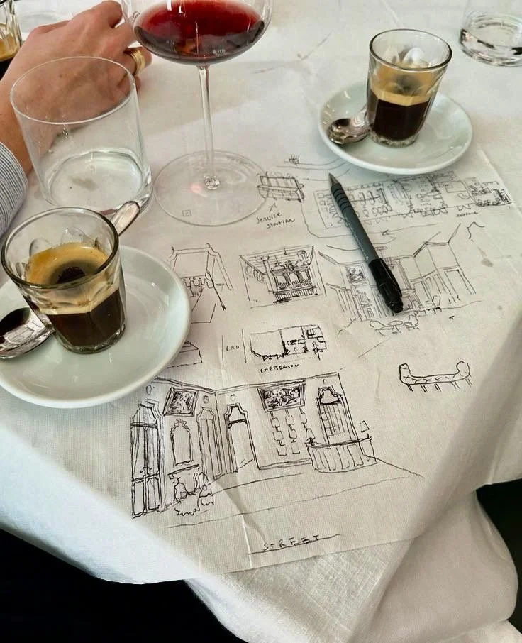 A table with three glasses of dark and clear liquids, a glass of red wine, a pen, and a sheet of paper with architectural sketches.