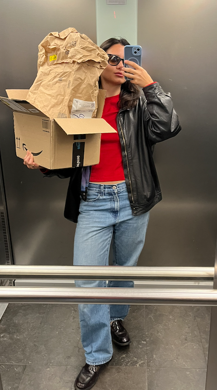 Woman wearing sunglasses, a black leather jacket, red shirt, and blue jeans, taking a mirror selfie in an elevator while holding a cardboard box filled with crumpled brown paper and a small package.