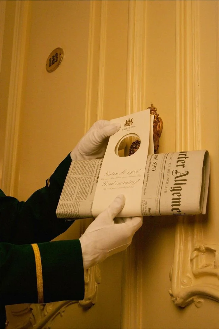 A person wearing white gloves handling a rolled-up newspaper, with a small card that says 'Guten Morgen! Good morning!' held by another gloved hand, in an ornate, classical interior setting.