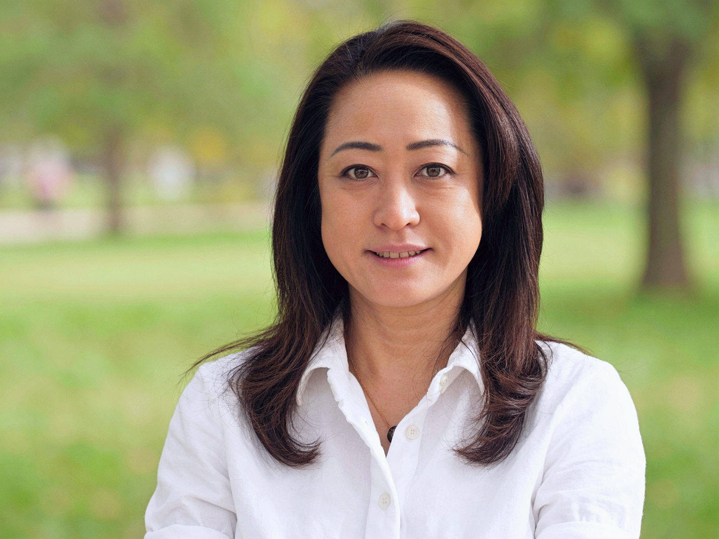 Portrait of an Asian woman with medium-length dark hair, smiling, outdoors in a park with green grass and trees in the background.