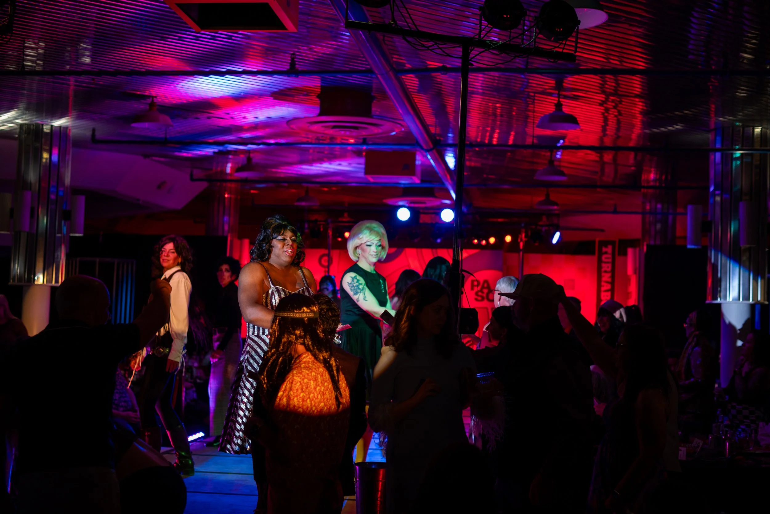Scene from a nightclub or event space with colorful lighting and people dancing and socializing. Two women are on stage, one with dark curly hair and striped dress, another with blonde short hair and tattoos, appear to be hosting or performing.
