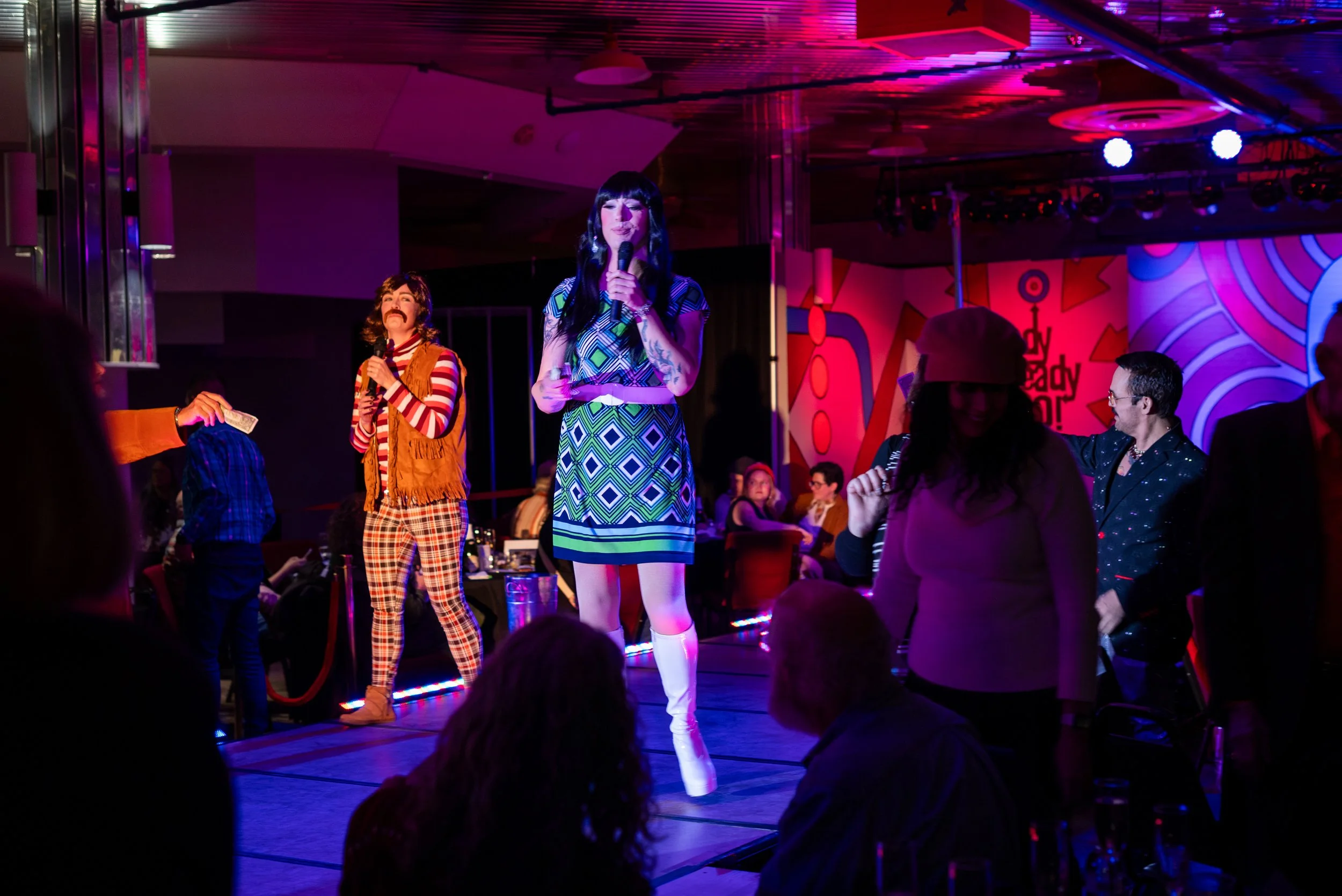 A drag queen performs on stage with a microphone, wearing a colorful geometric dress and white thigh-high boots. The background features vibrant red and purple abstract graffiti art, with party guests seated and standing around the stage, enjoying th