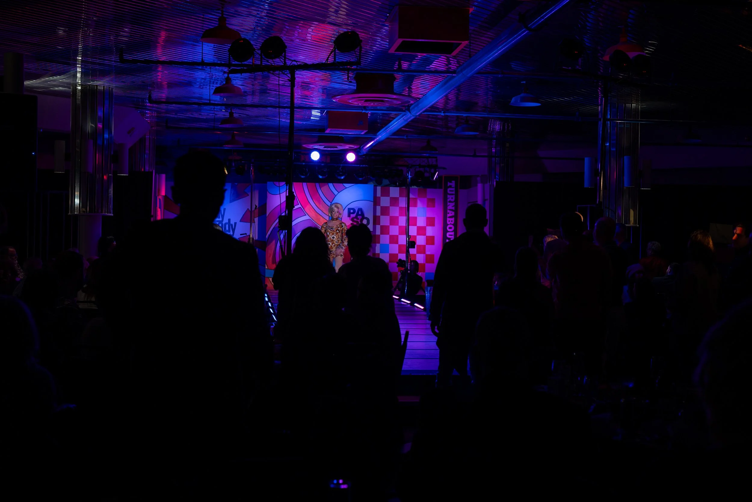 A performer on stage at a comedy or variety show, with an audience watching in a dimly lit venue with colorful stage lighting and background decorations.