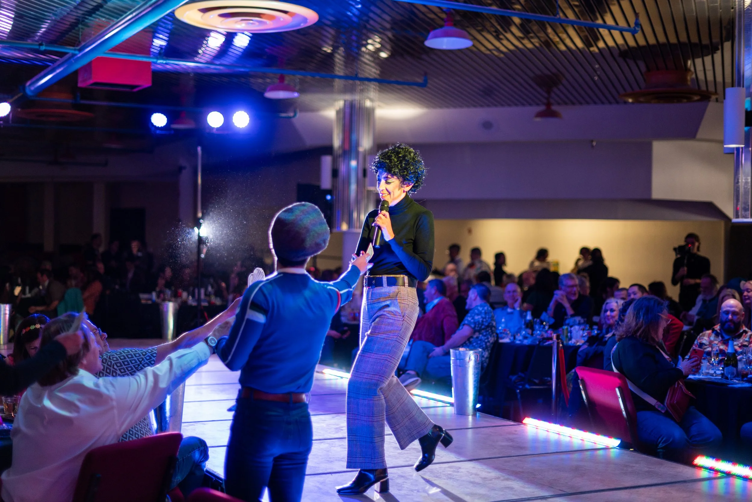 A woman with curly hair holding a microphone on stage, interacting with a child reaching out to her, in a dimly lit venue filled with seated audience members watching her perform.