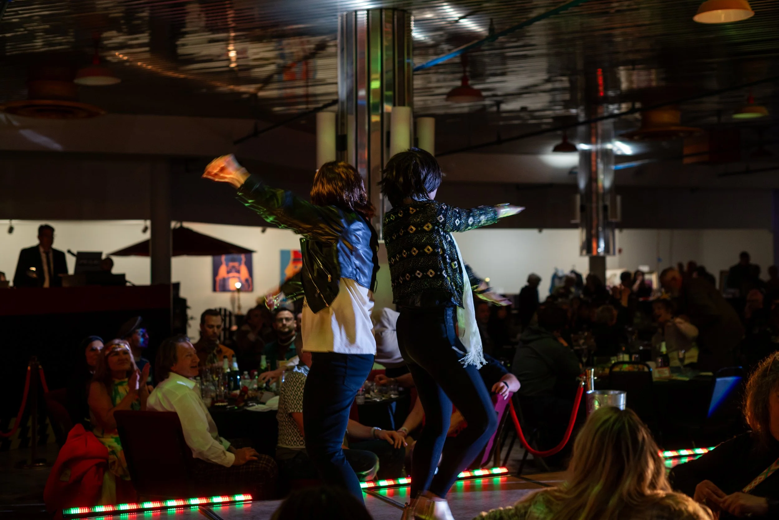 People dancing on a stage in front of seated audience in a dimly lit room, with colorful lights.