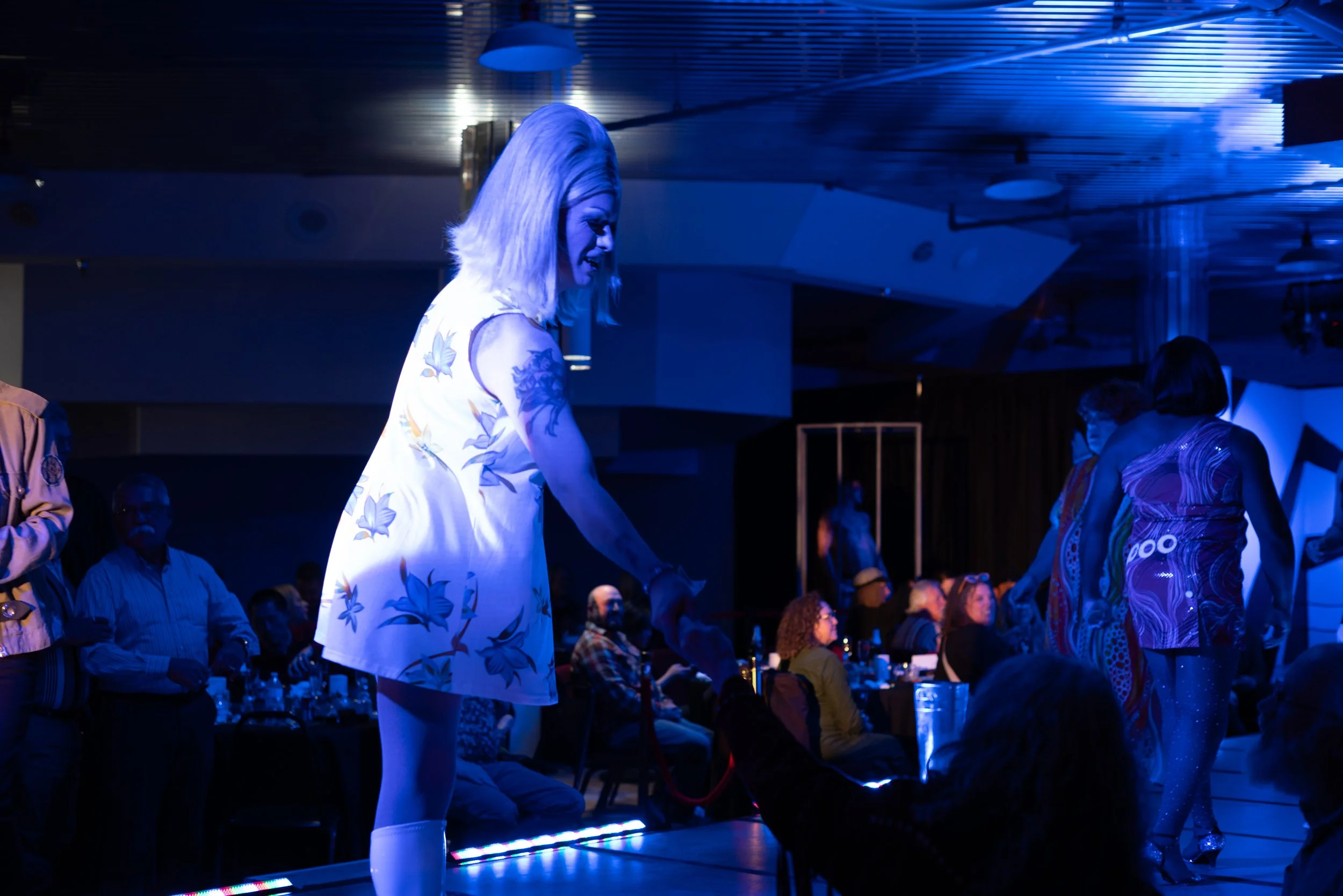 A woman with blonde hair, tattoos on her arms, and wearing a white dress with blue floral patterns is standing on a stage, illuminated by blue stage lighting, in a crowded indoor venue with an audience seated and watching her perform or speak.