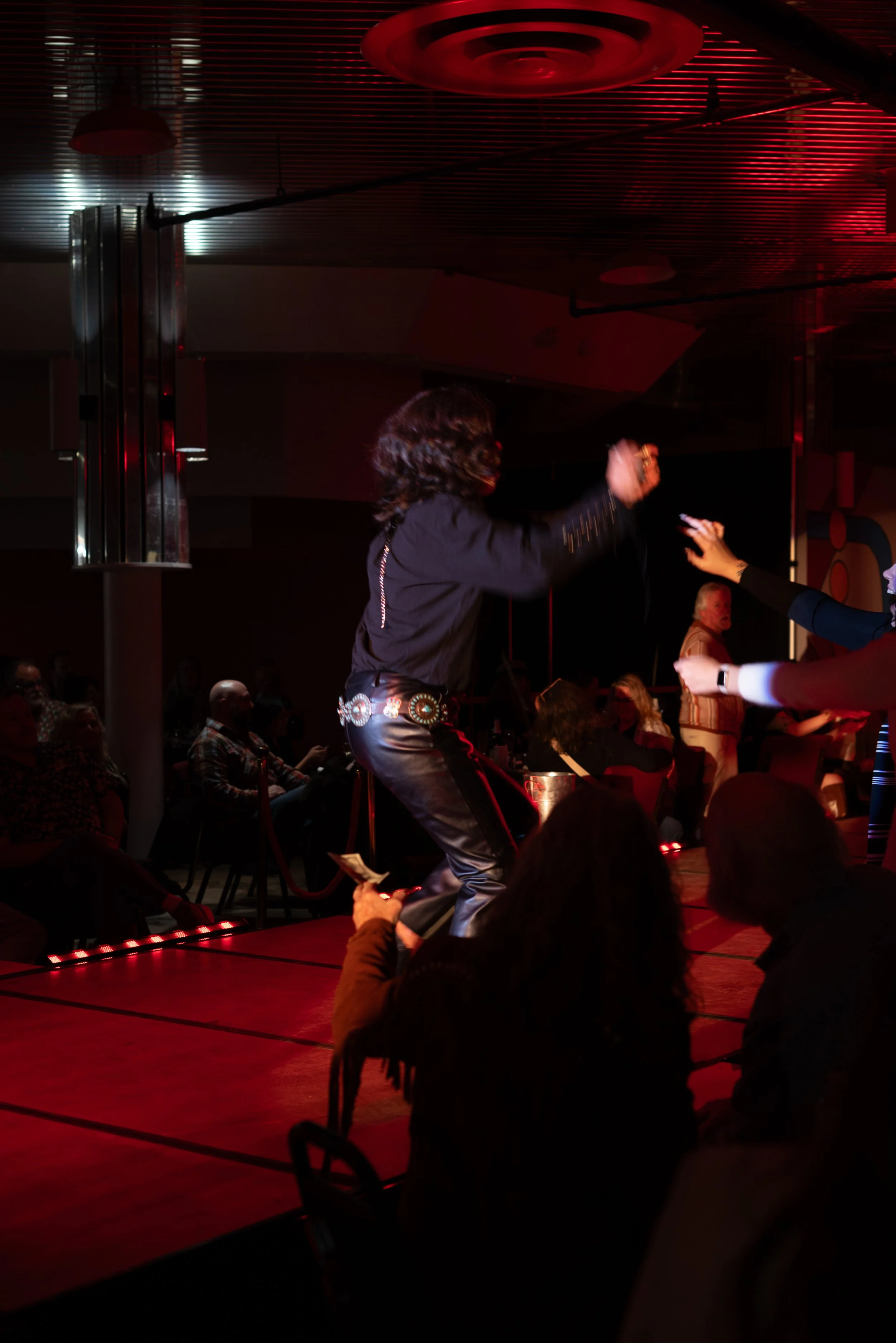 A person with curly hair is dancing or singing on a stage at an indoor event with red lighting, surrounded by an audience.