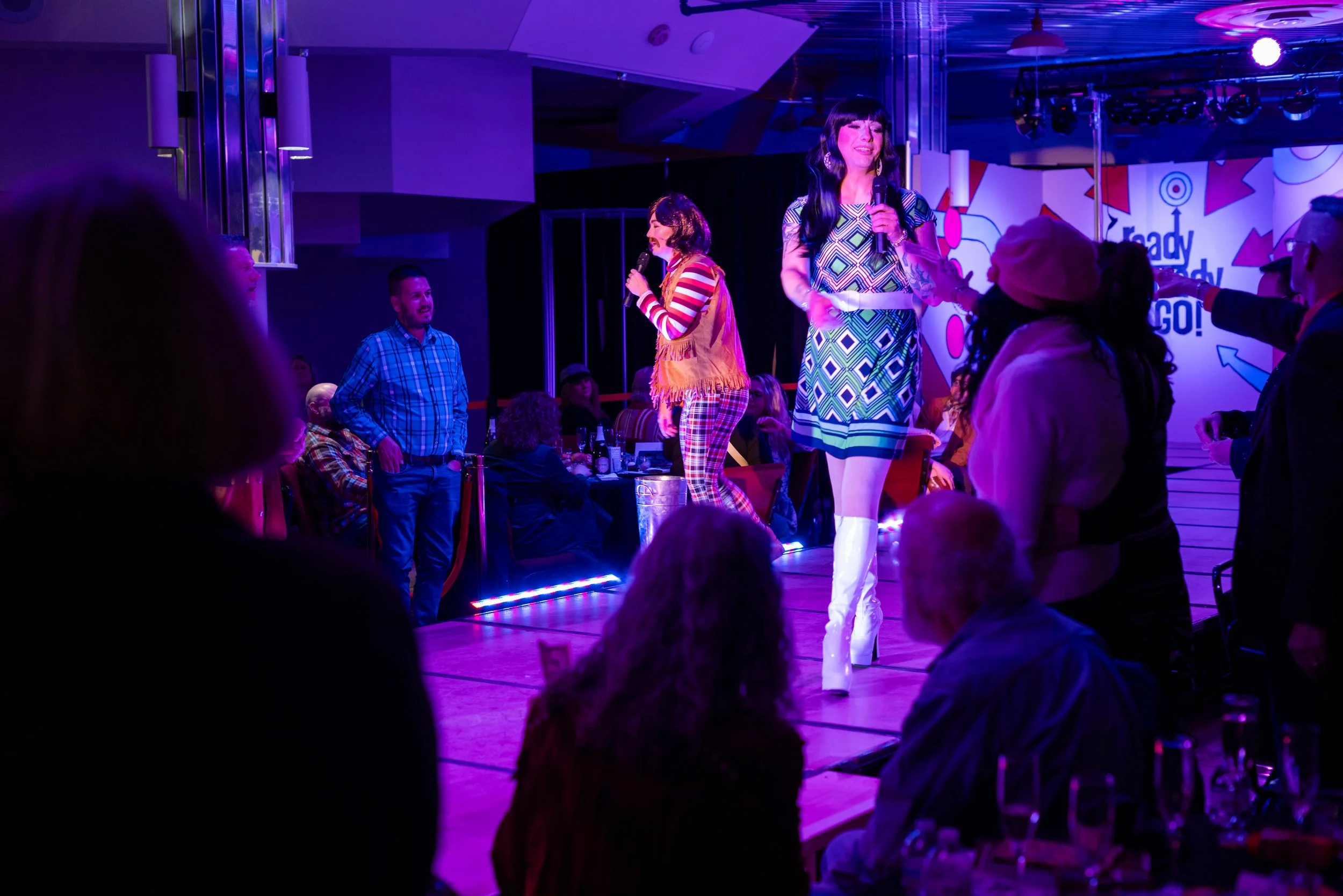 People on stage performing at a lively event with vibrant purple and pink lighting, audience members watching and enjoying the show.