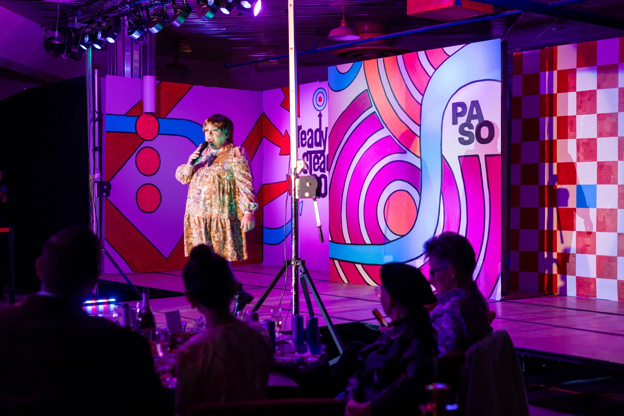 A woman in a patterned dress performs on a stage with colorful, abstract background art, called "Ready Steady Go!". Audience members are seated at tables watching her in a dimly lit venue.