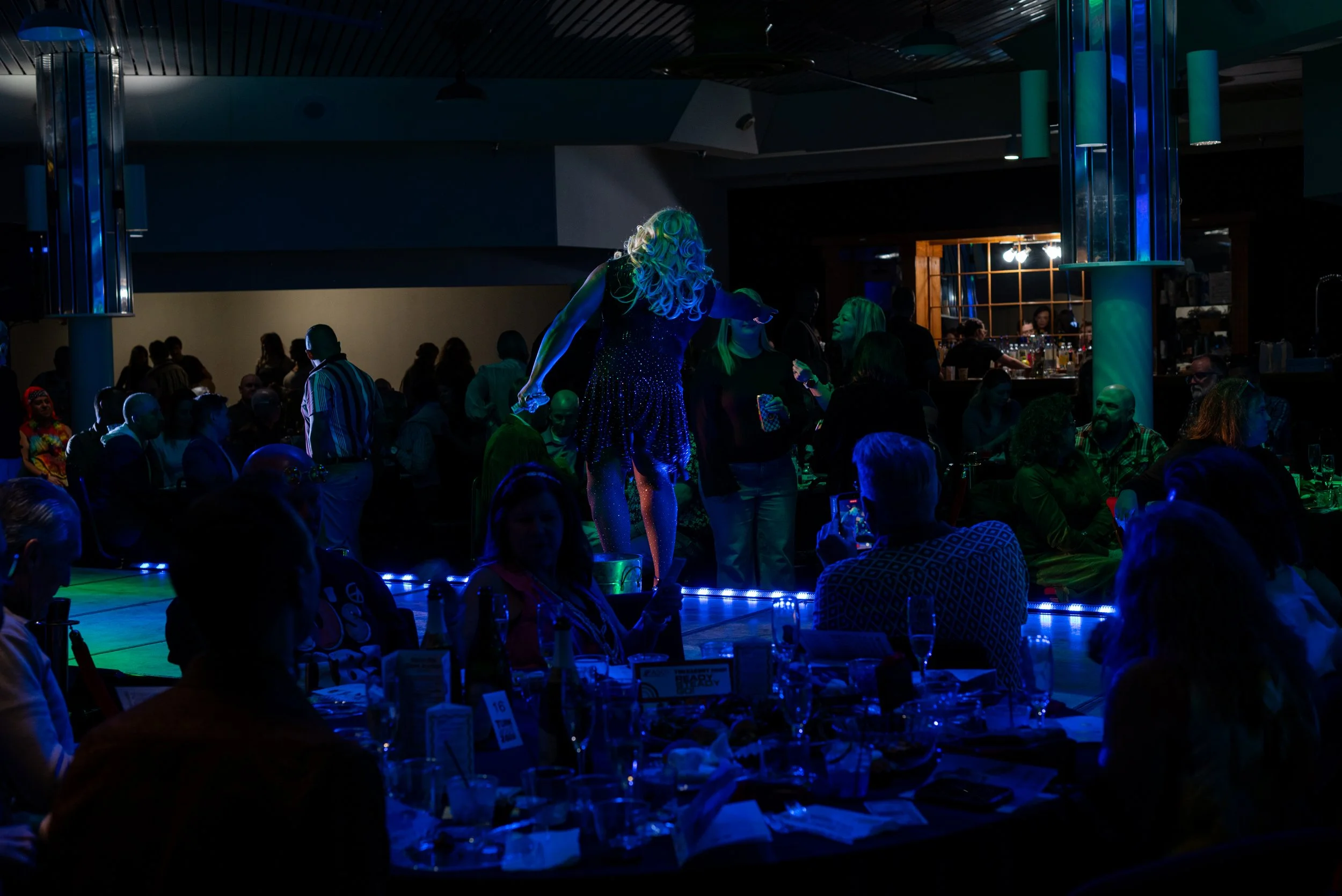Couples and groups of people seated around tables at a nightlife event with a performer in a glittery dress on a small stage, illuminated by blue and green stage lighting.