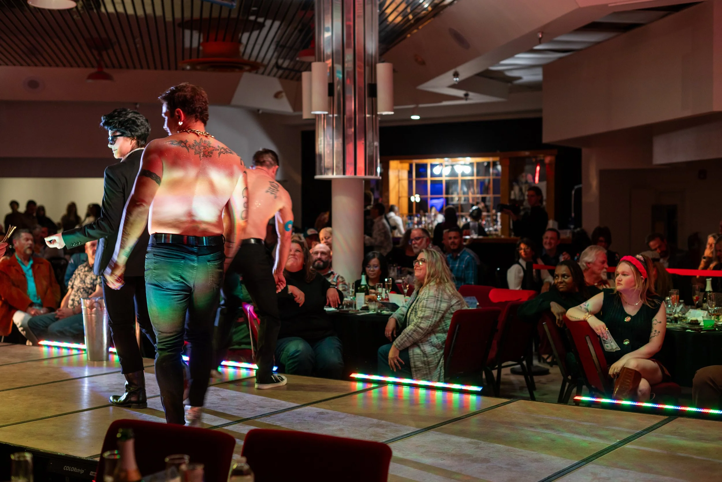 People watching a drag show on stage at a nightclub or bar, with some performers on stage and an audience seated at tables, decorated with colored lighting.