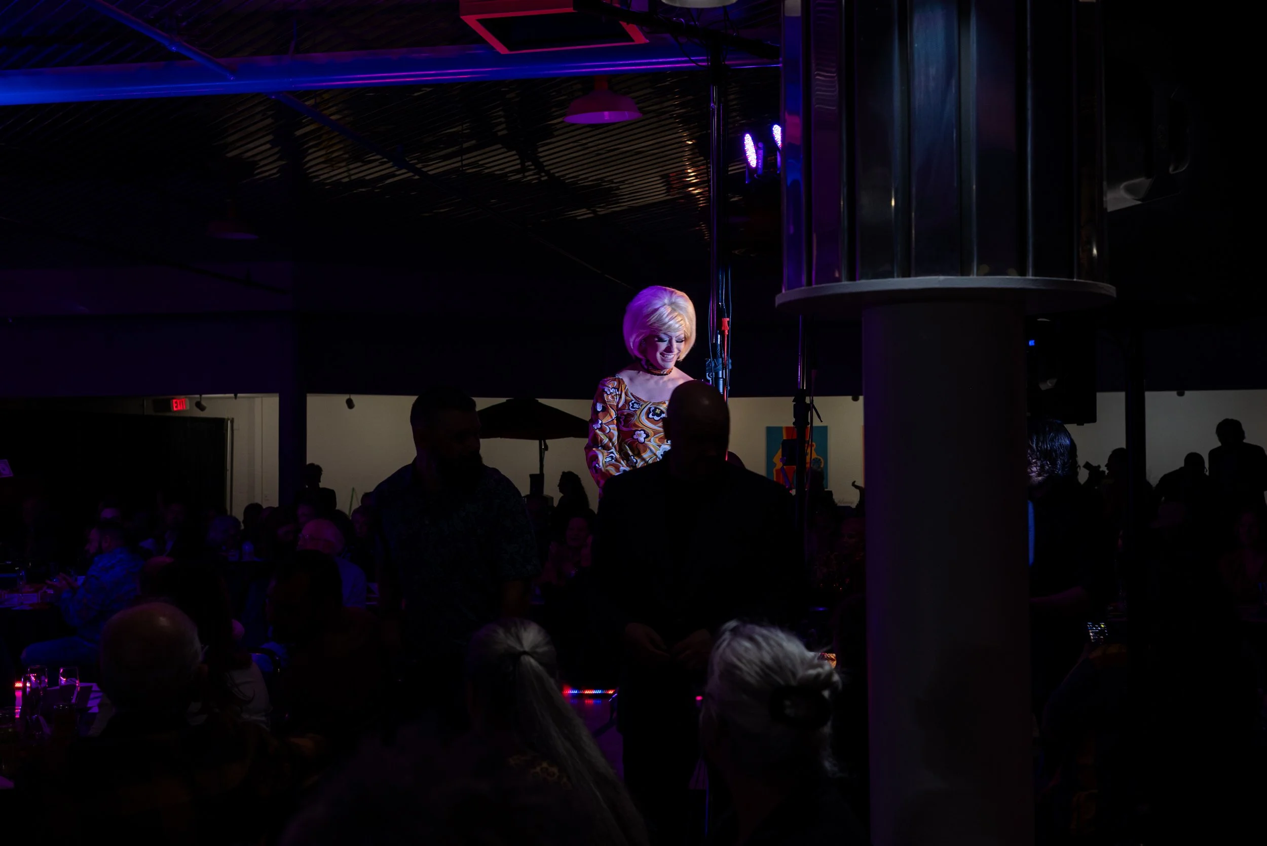 A woman with blonde hair on stage, smiling, in a colorful patterned top. Audience members are seated in a darkened venue with purple and blue lighting.