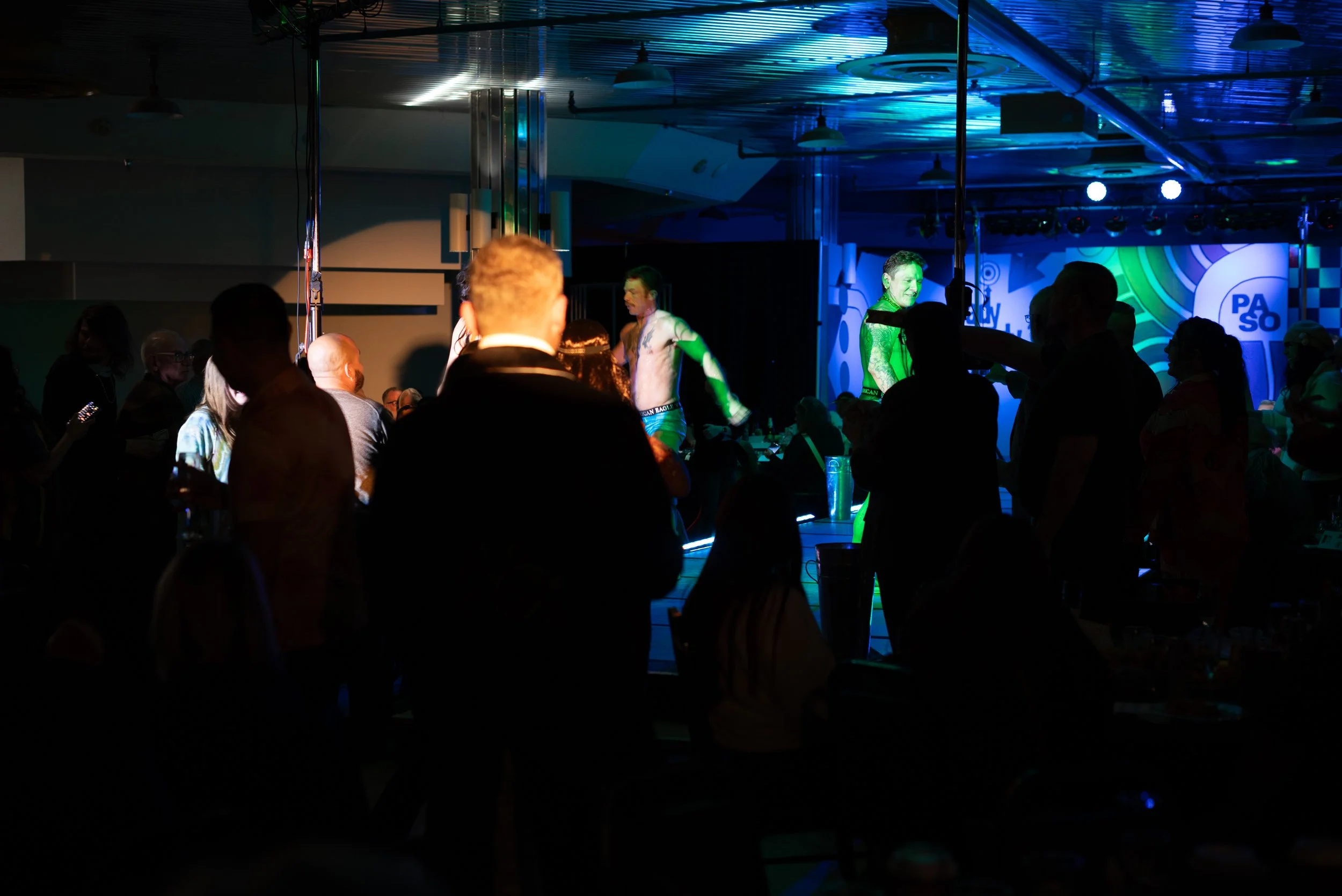 A lively event with people watching a performance on stage in a dark, colorful room. The stage has bright lighting and a background displaying the PASO logo, with performers including a shirtless man and a woman with vibrant green hair.