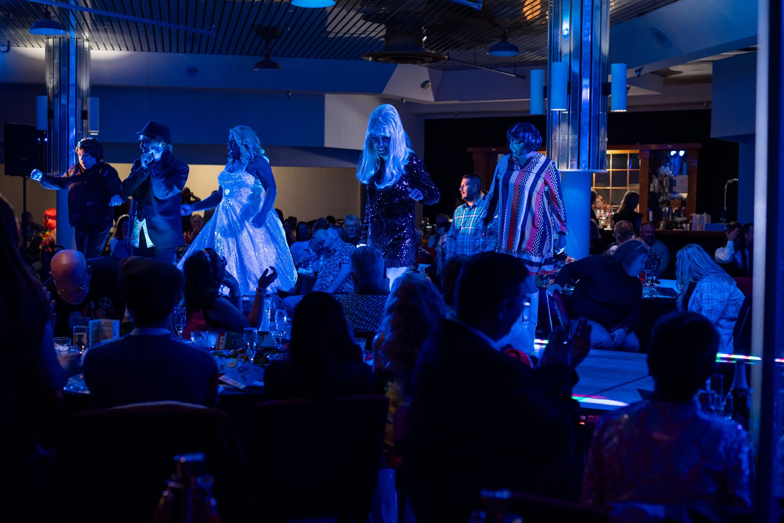 Performance at a lively event with multiple performers dressed in colorful, sparkly outfits under blue lighting, audience seated at tables watching.