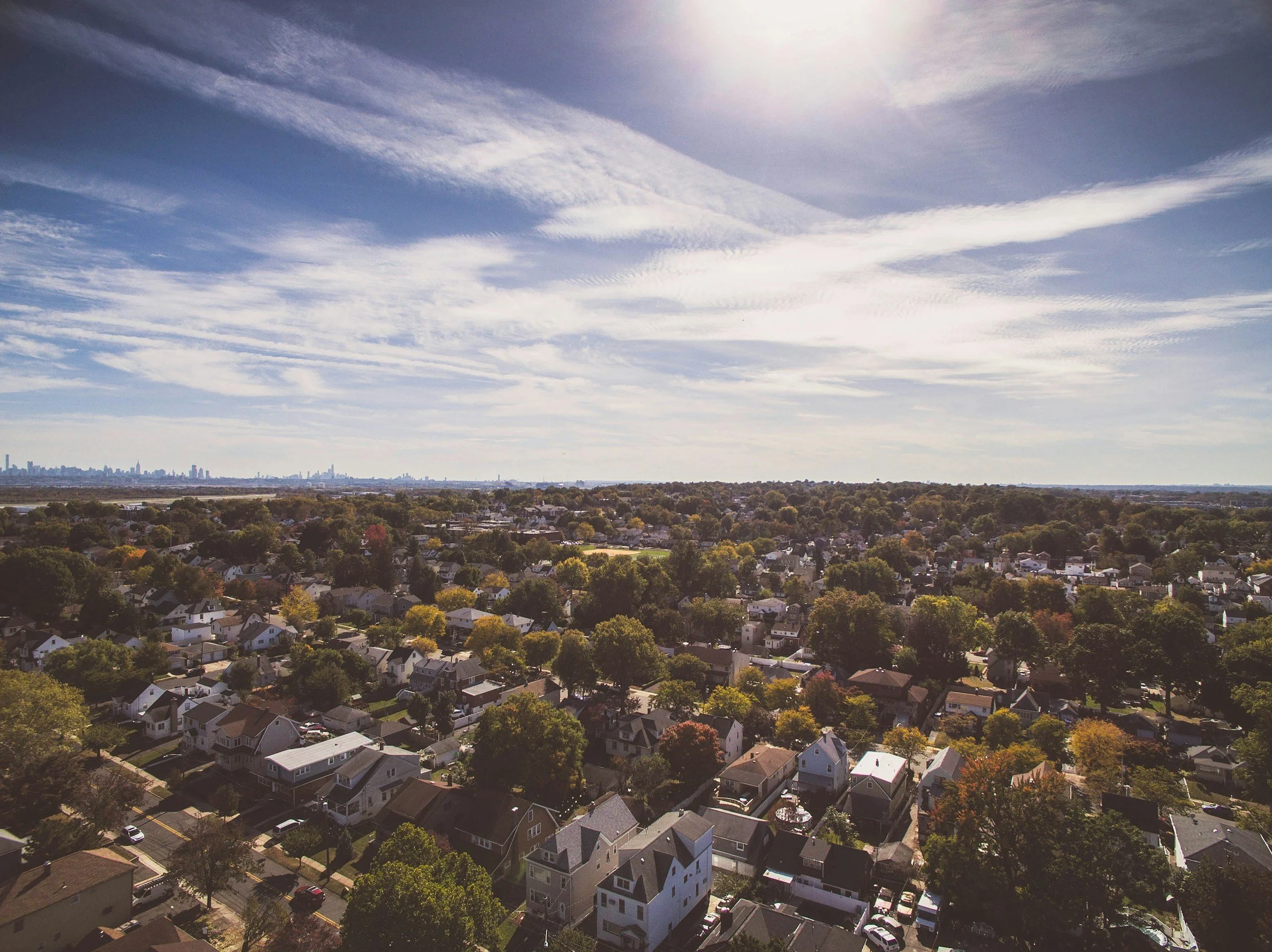 Aerial view of a suburban neighborhood with trees, houses, and streets, under a partly cloudy sky with sunlight.
