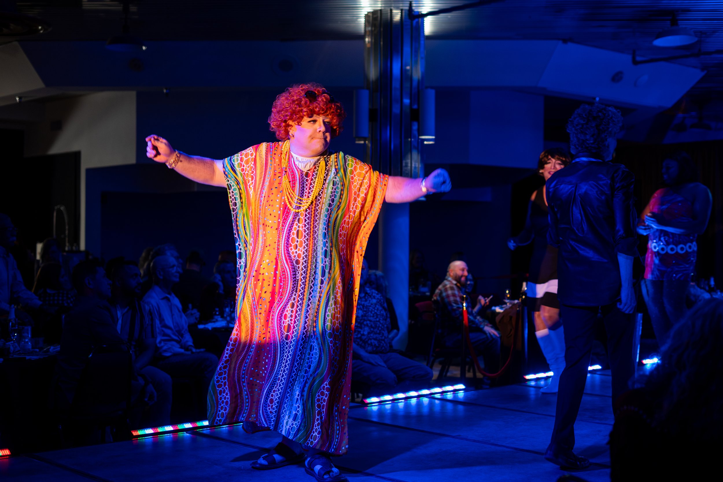 A performer dressed in a colorful, flowing dress with a curly red wig, sunglasses, and jewelry, dancing on stage at a lively event with people seated and standing around.