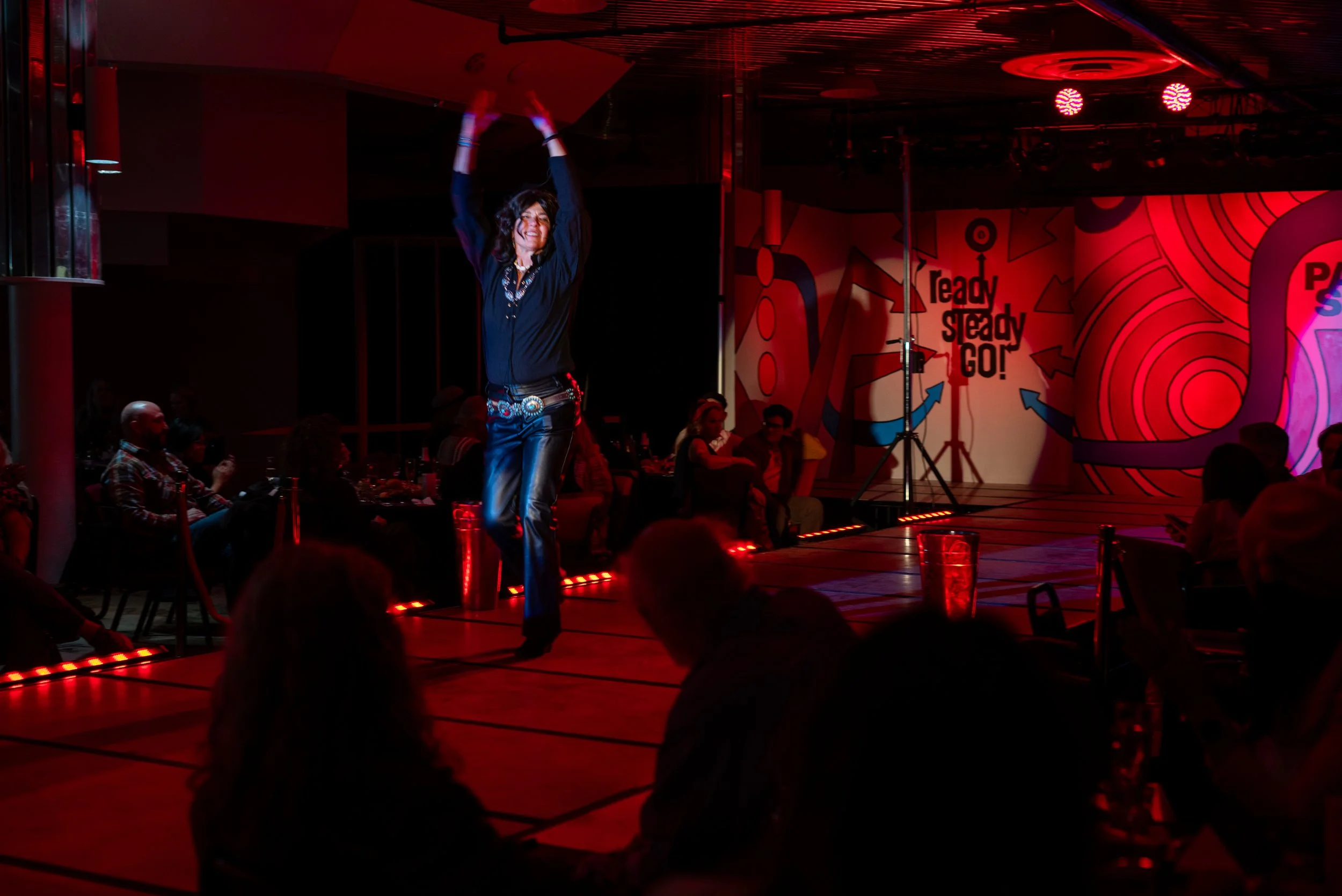A woman dancing on a stage with red and purple lighting, wearing black pants and a black top, with a crowd watching her.