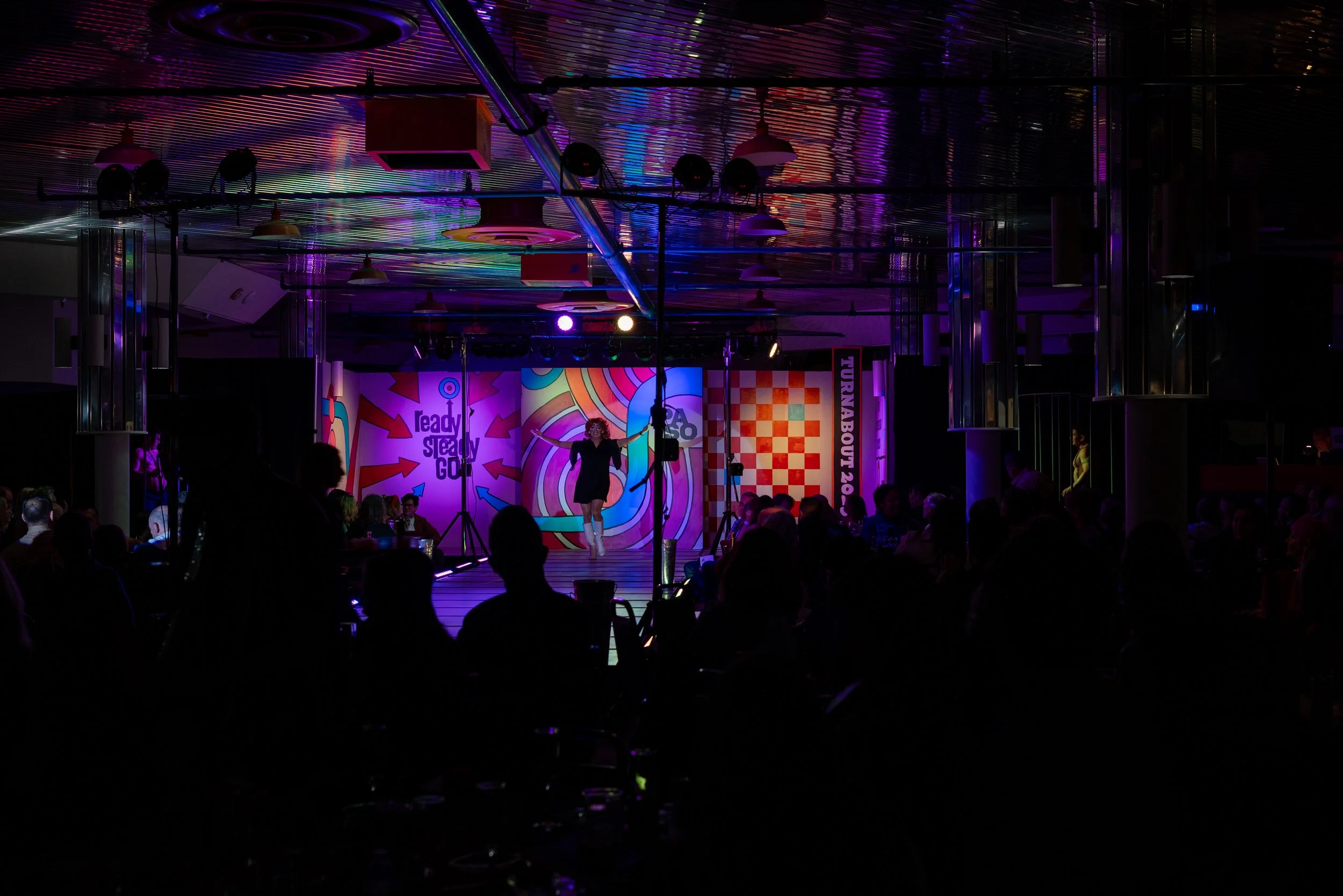 A performer on stage at an event with colorful, abstract background and audience watching inside a dimly lit venue.