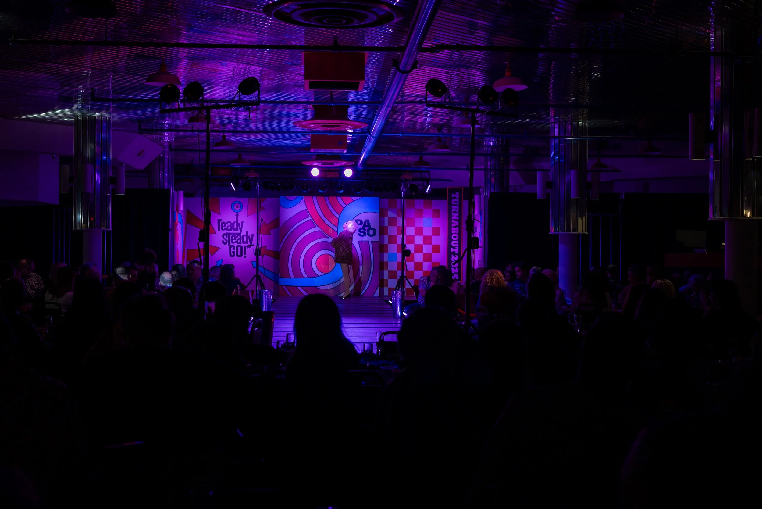 Stage with colorful graphics and text, performer dressed in a patterned outfit, audience sitting in front under purple lighting, at a live event or show.