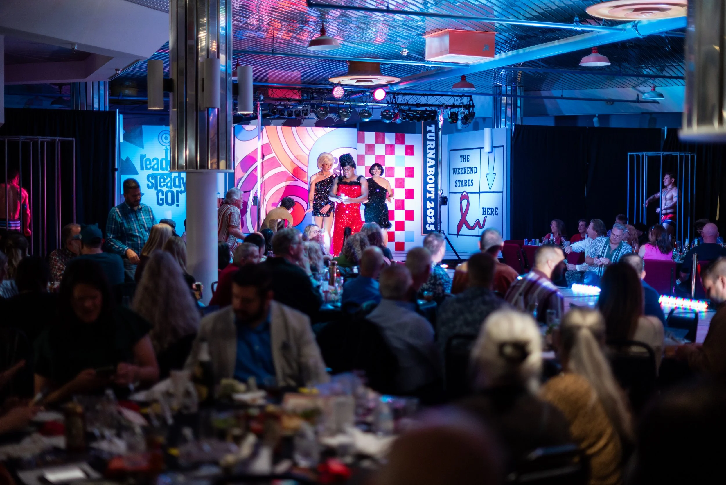 People attending an indoor event watching a stage performance with performers dressed in retro outfits, illuminated by colorful stage lighting, with signs indicating a themed weekend event.