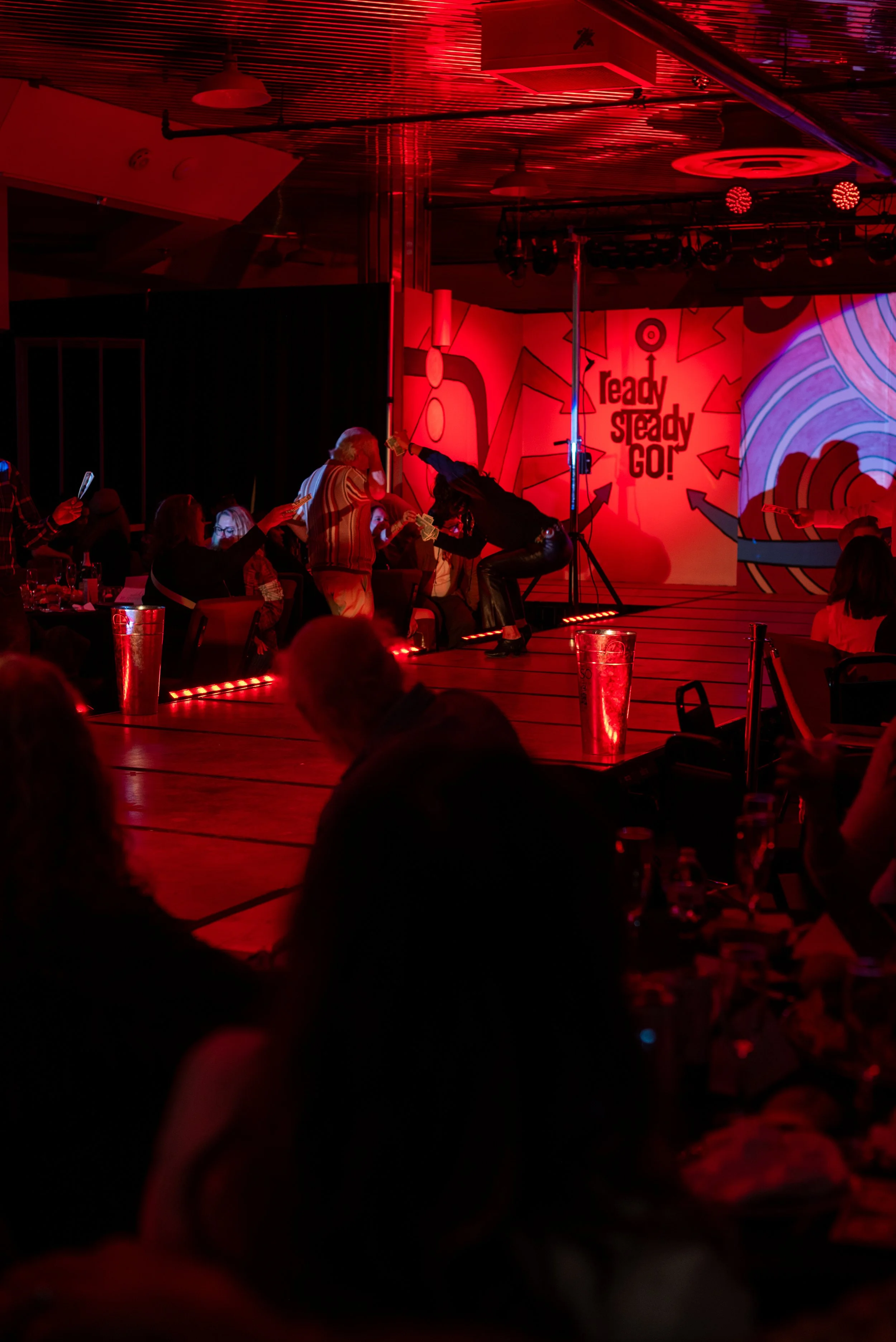 A stage with a red and pink abstract background that has the words "Ready, Steady, Go!" on it. There are performers on stage, including a man and a woman, with the woman leaning forward as she dances or performs. Audience members are seated at tables