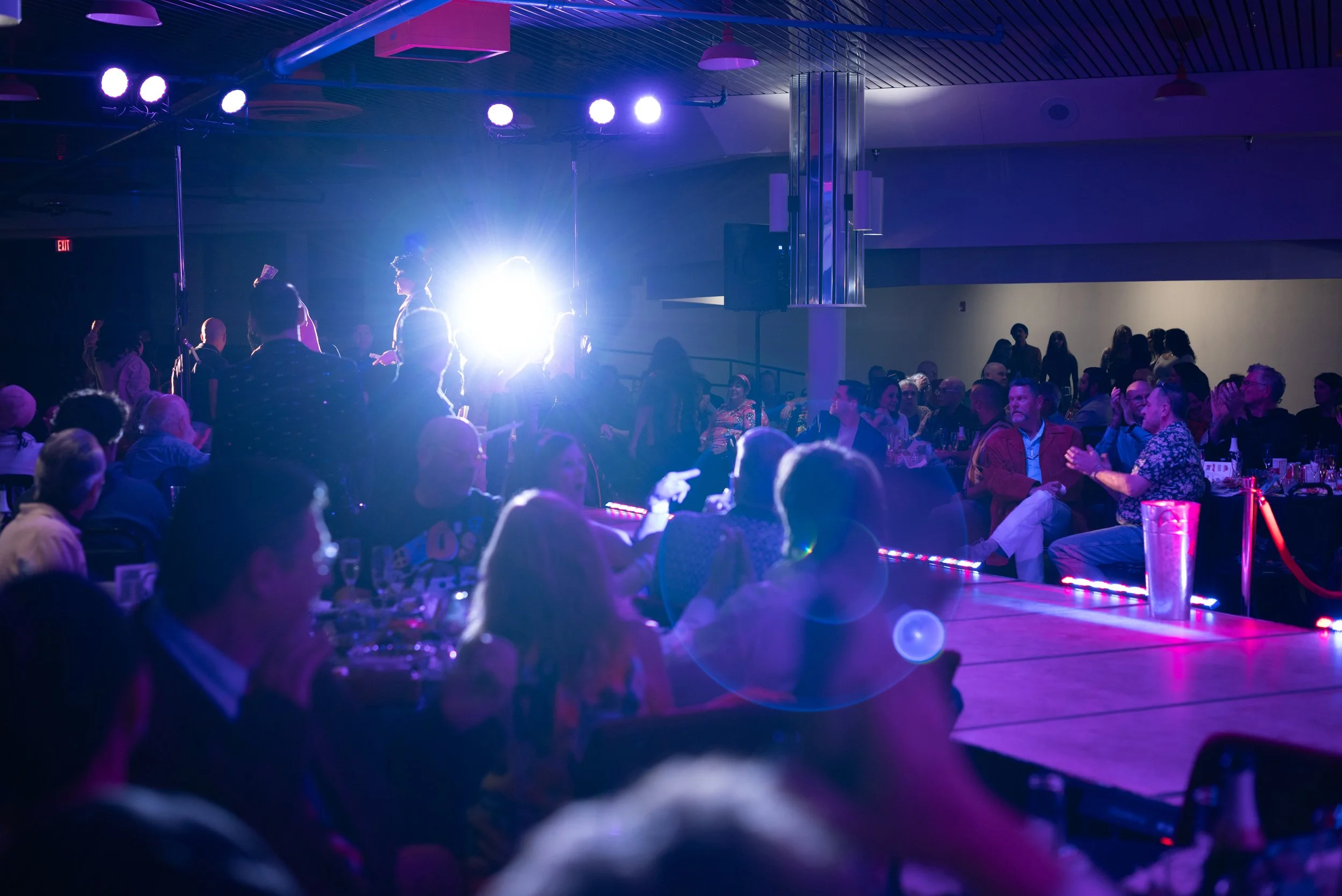 A lively indoor event with a stage, purple and blue lighting, and an audience seated at tables, watching a performance or presentation.