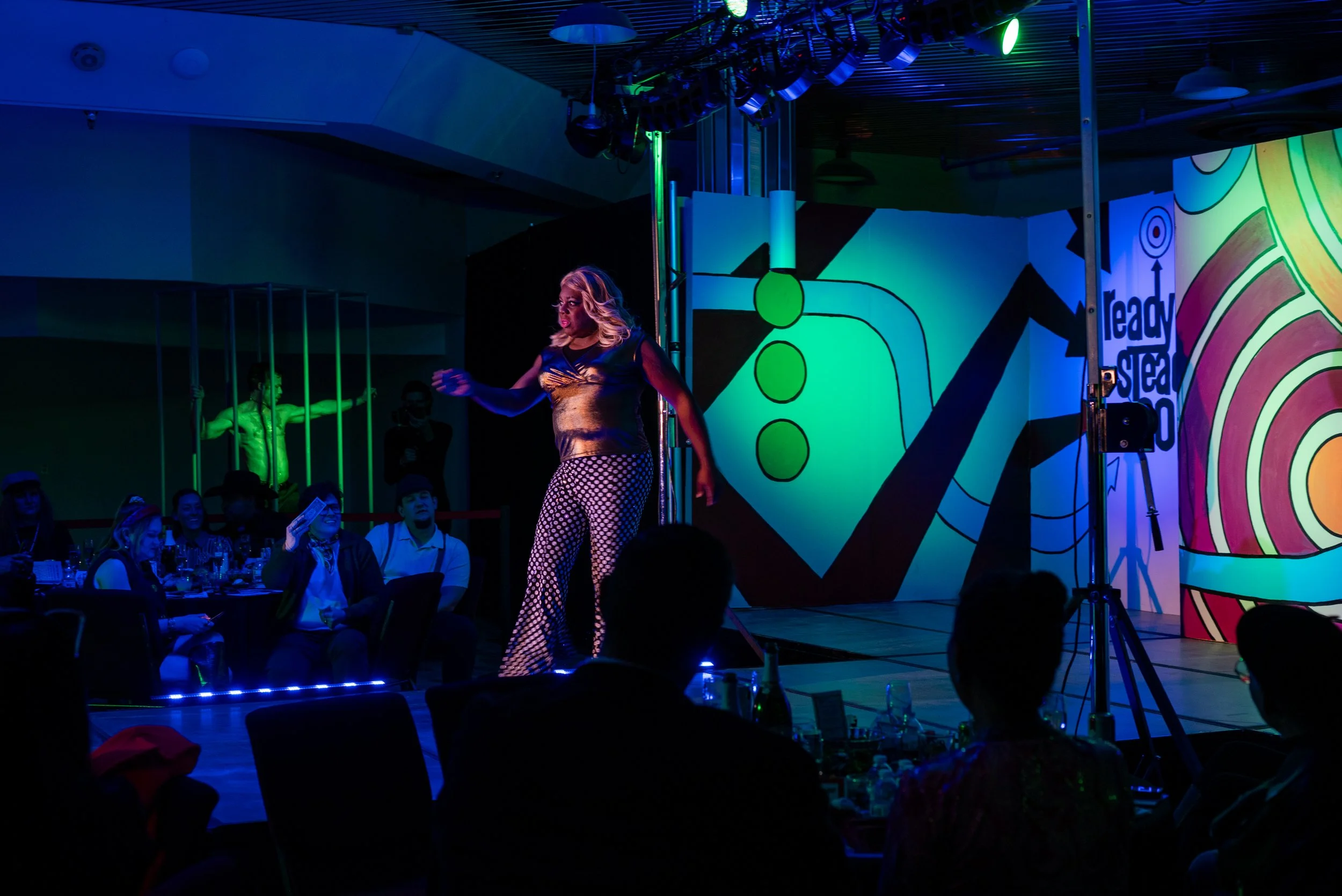 Performer on stage at a lively entertainment event, with colorful abstract background and audience seated at tables.