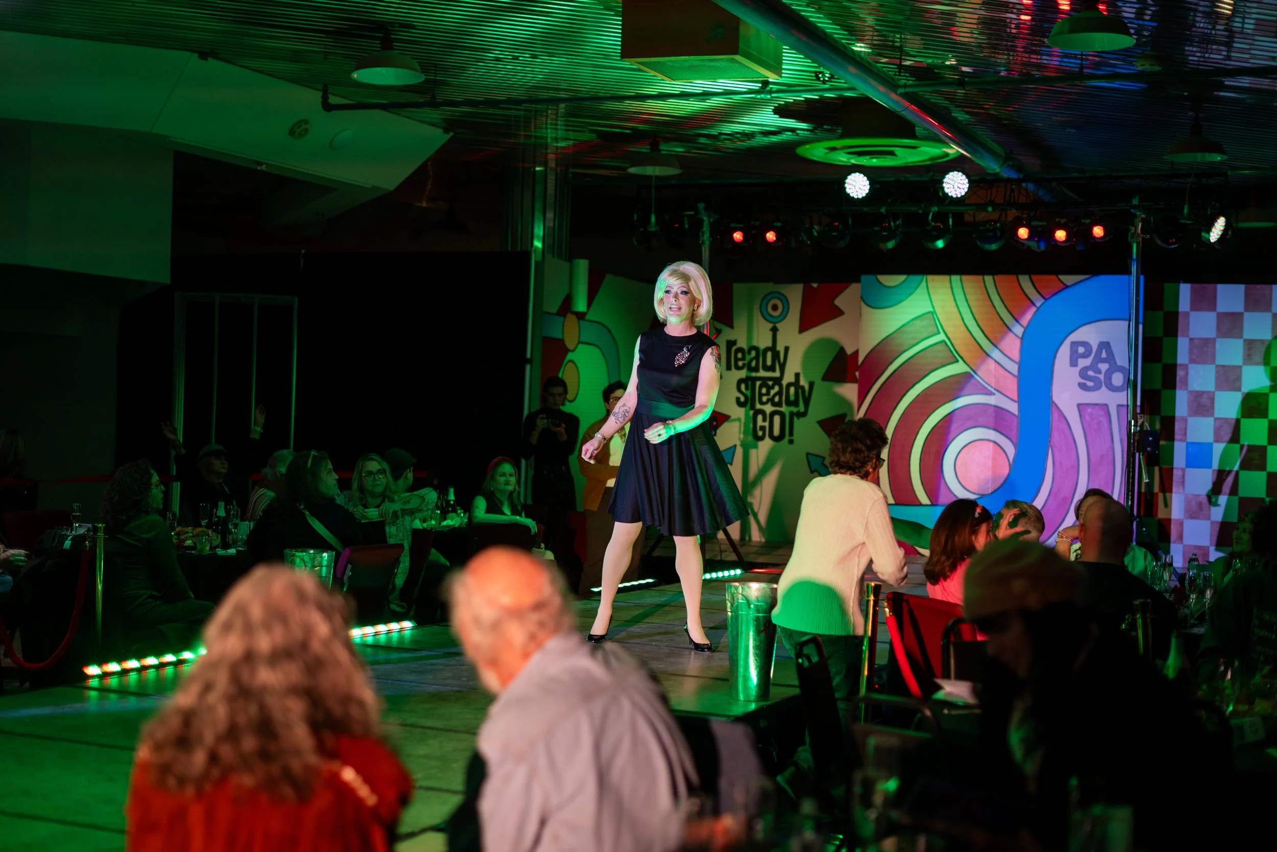 A woman with blonde hair performing on a stage at a lively event. She is wearing a black dress with a brooch and heels, standing in front of a colorful, abstract background with the text 'Ready Steady GO!' The audience is seated at tables, watching h