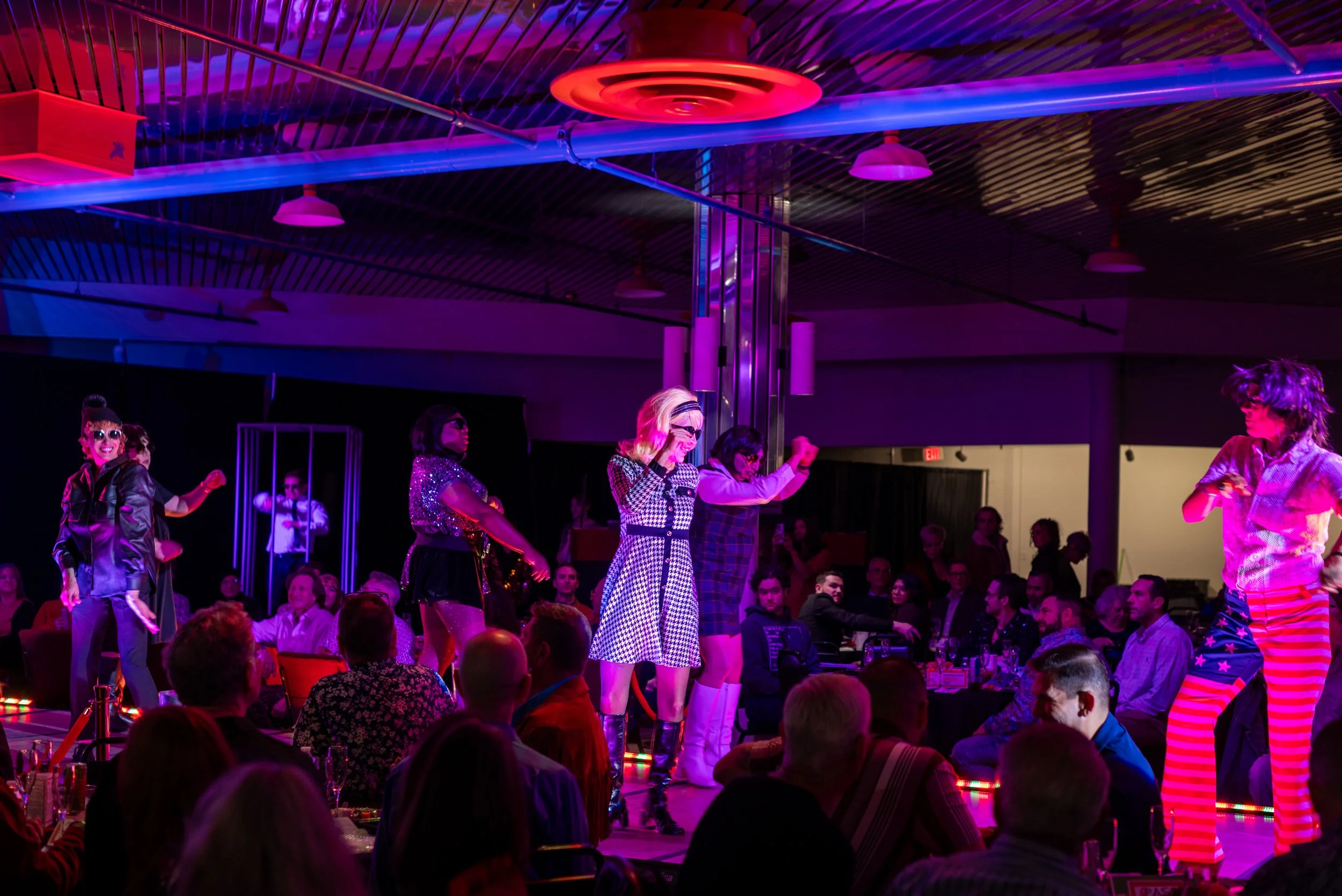Performers dancing on stage at a nightclub with an audience seated at tables, illuminated by colorful stage lights.
